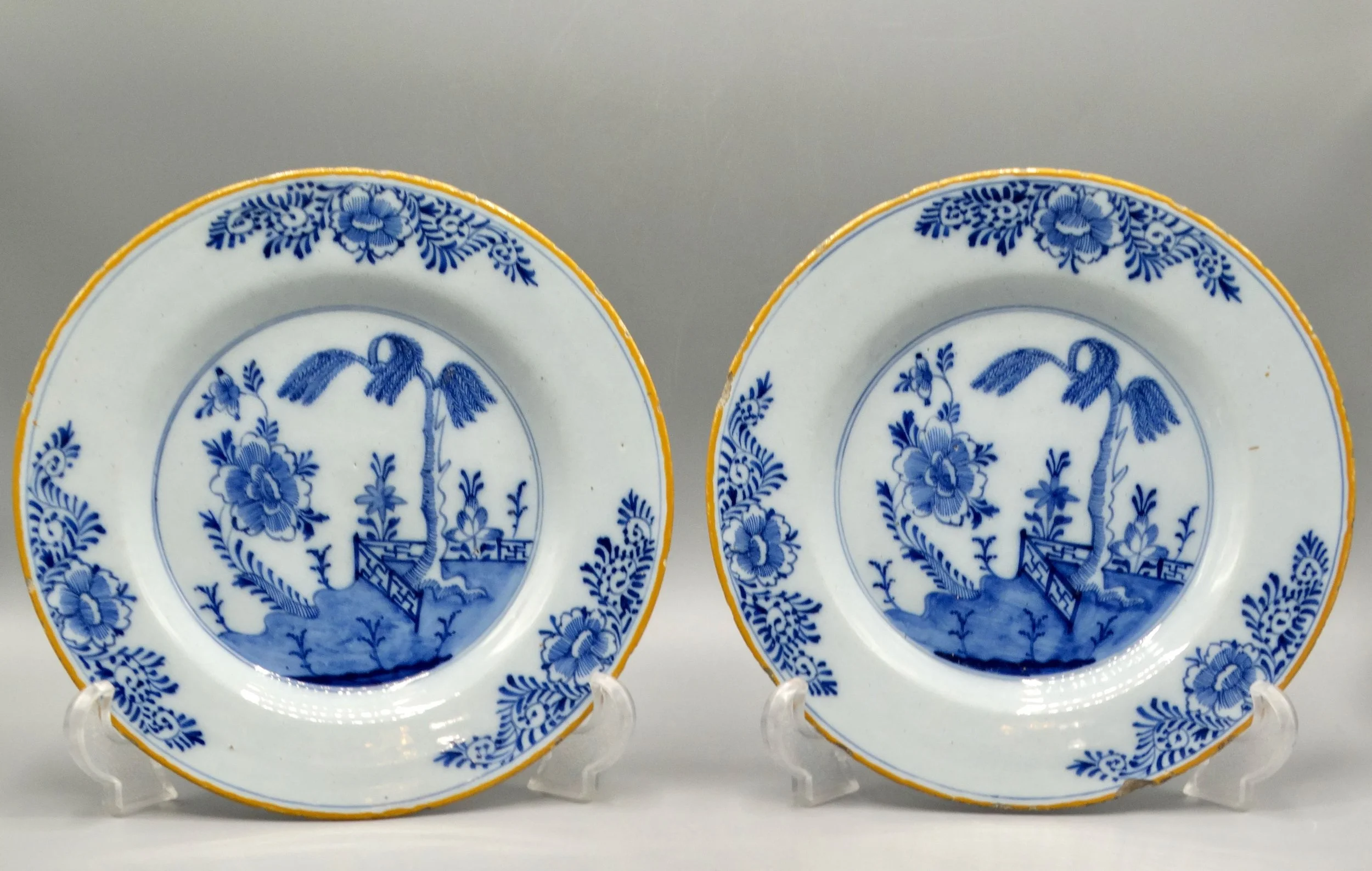 Pair of 18th-century Dutch Delft faience plates with chinoiserie tree, fence, floral and geometric motifs, orange-yellow borders.