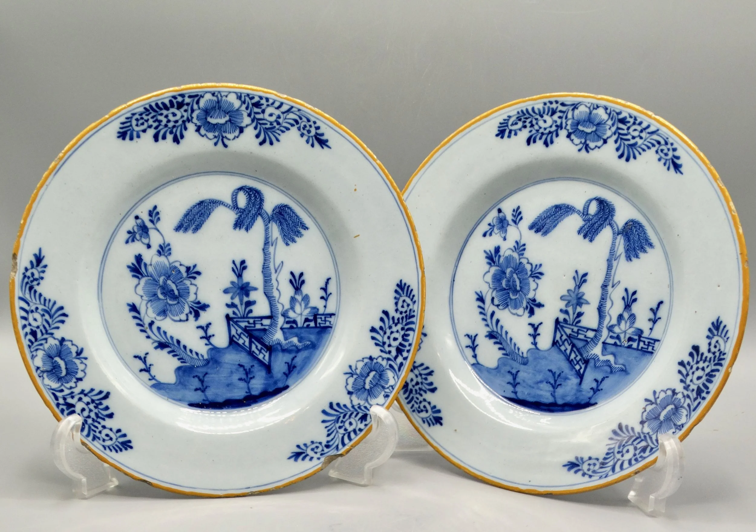 Pair of 18th-century Dutch Delft faience plates with chinoiserie tree, fence, floral and geometric motifs, orange-yellow borders.