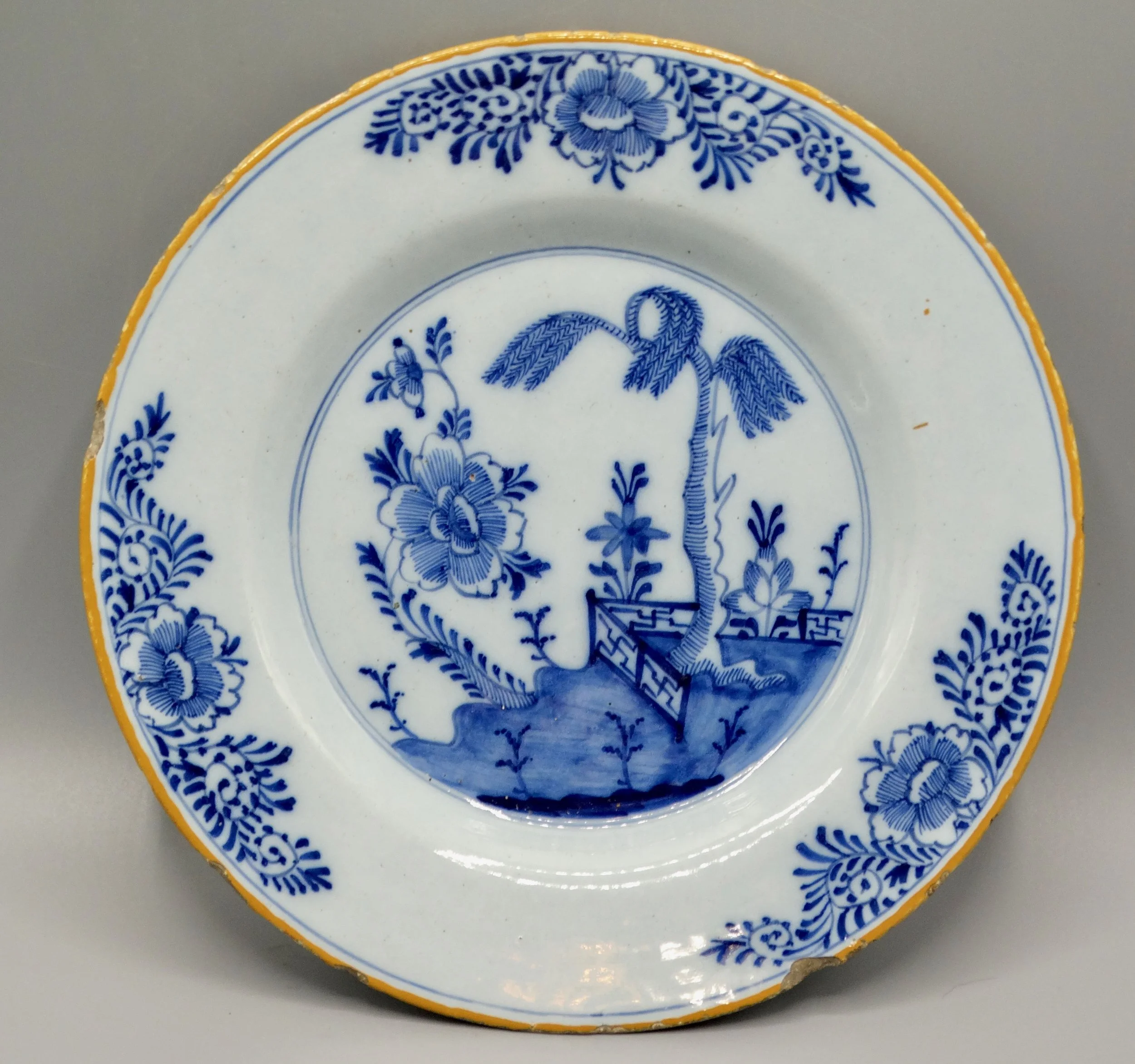 18th-century Dutch Delft faience plate with chinoiserie tree, fence, floral and geometric motifs, orange-yellow border.