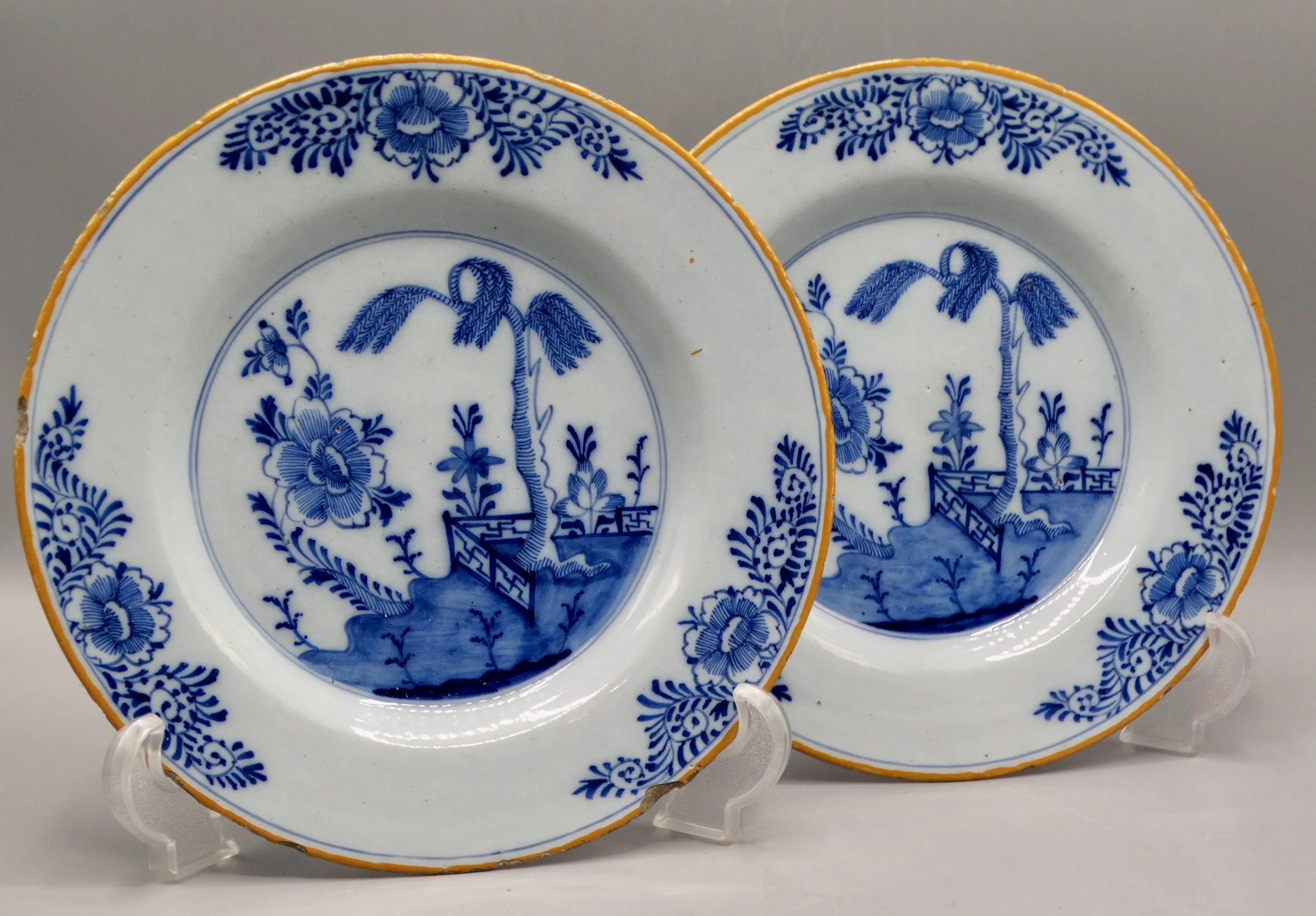 Pair of 18th-century Dutch Delft faience plates with chinoiserie tree, fence, floral and geometric motifs, orange-yellow borders.