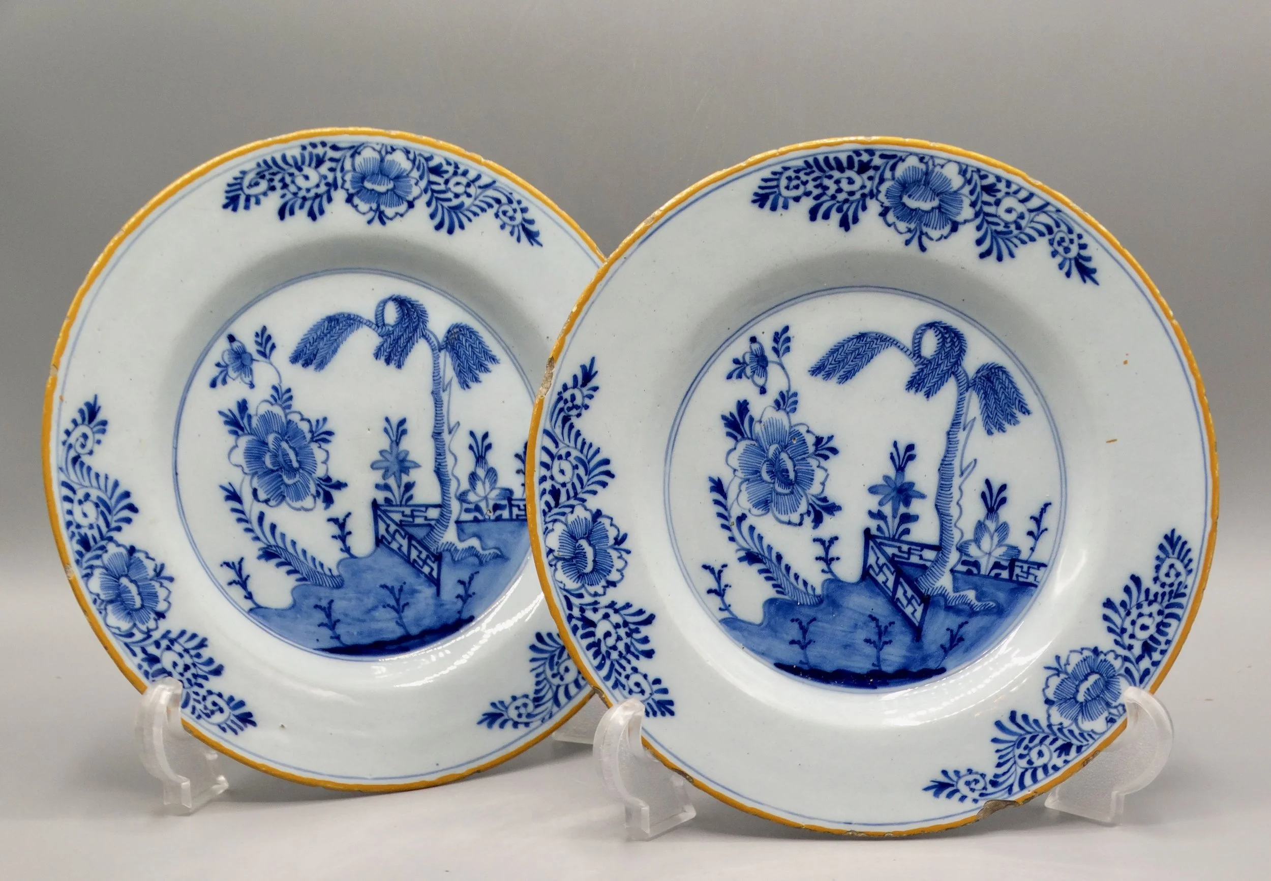 Pair of 18th-century Dutch Delft faience plates with chinoiserie tree, fence, floral and geometric motifs, orange-yellow borders.