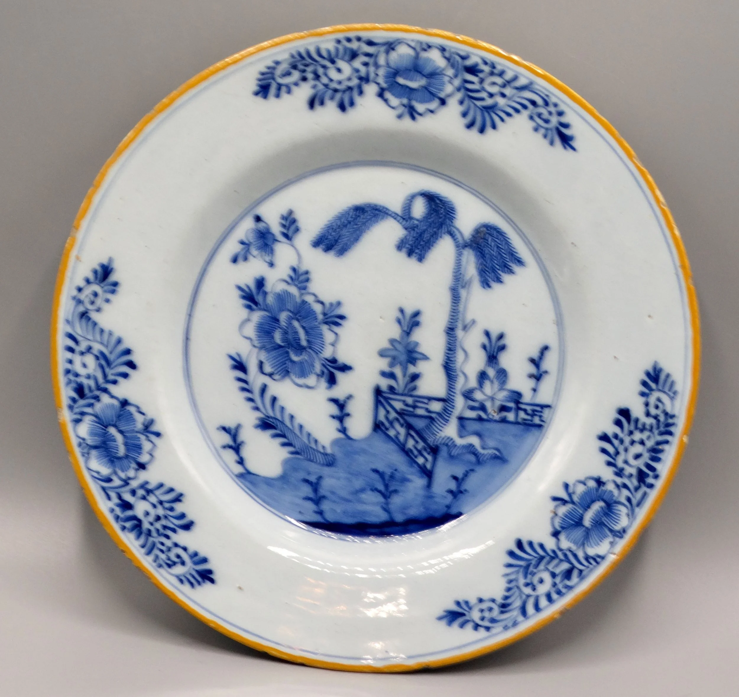Second 18th-century Dutch Delft faience plate with chinoiserie tree, fence, floral and geometric motifs, orange-yellow border.