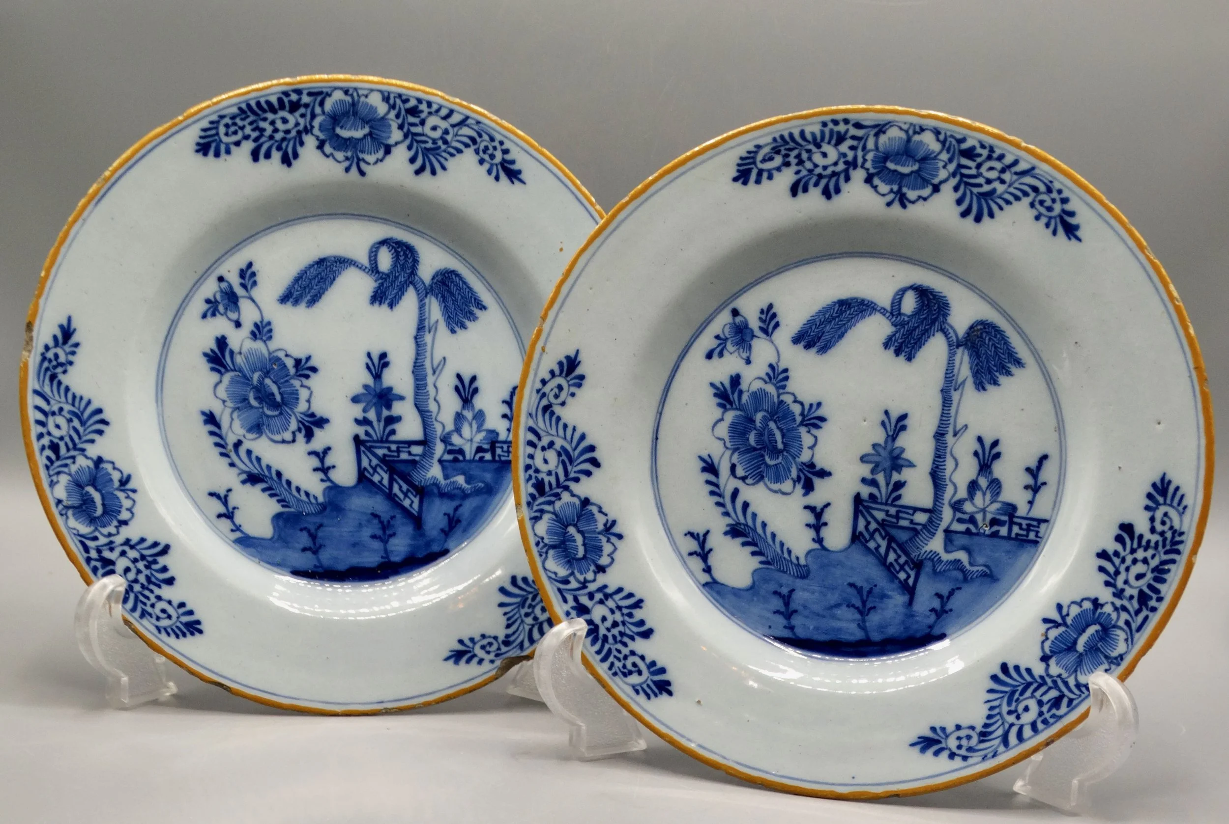 Pair of 18th-century Dutch Delft faience plates with chinoiserie tree, fence, floral and geometric motifs, orange-yellow borders.