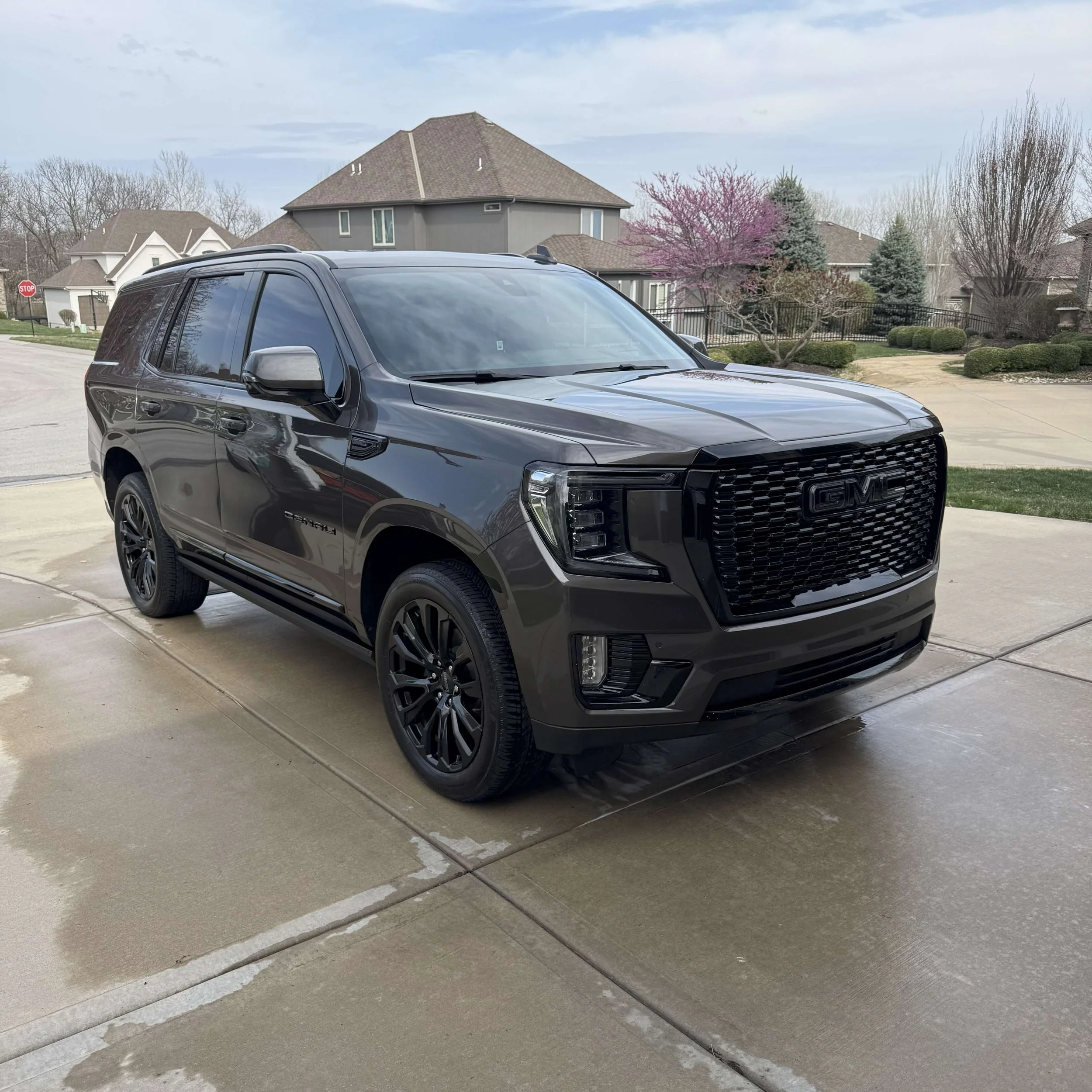 A black GMC SUV parked on a concrete driveway with a residential neighborhood background, including houses, trees, and a stop sign.