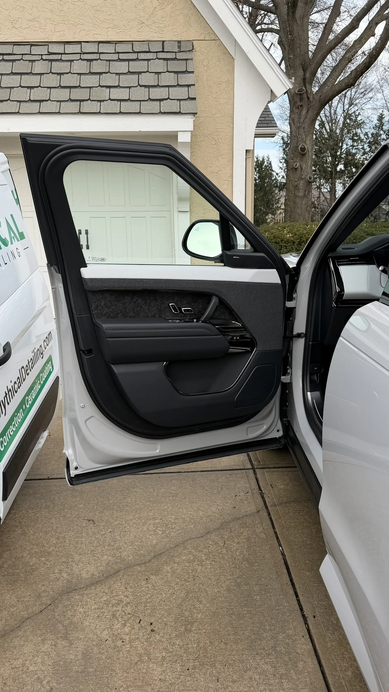 Open car door showing black and white interior with window controls, hinged against a white vehicle parked in a driveway with a beige house, garage, and leafless tree in the background.