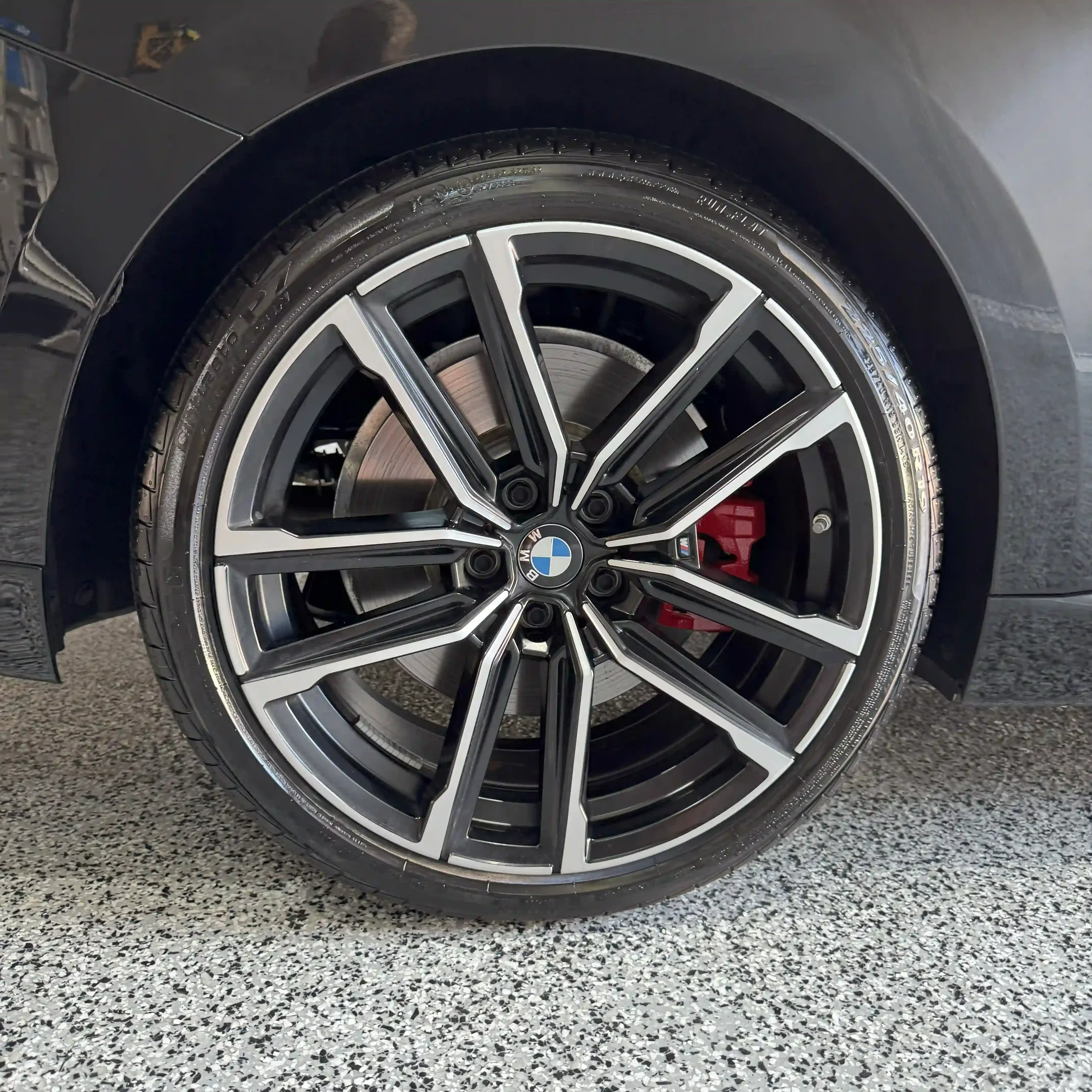 Close-up of a BMW car wheel with a black and silver rim, red brake caliper, and branded center cap, on a gray speckled garage floor.
