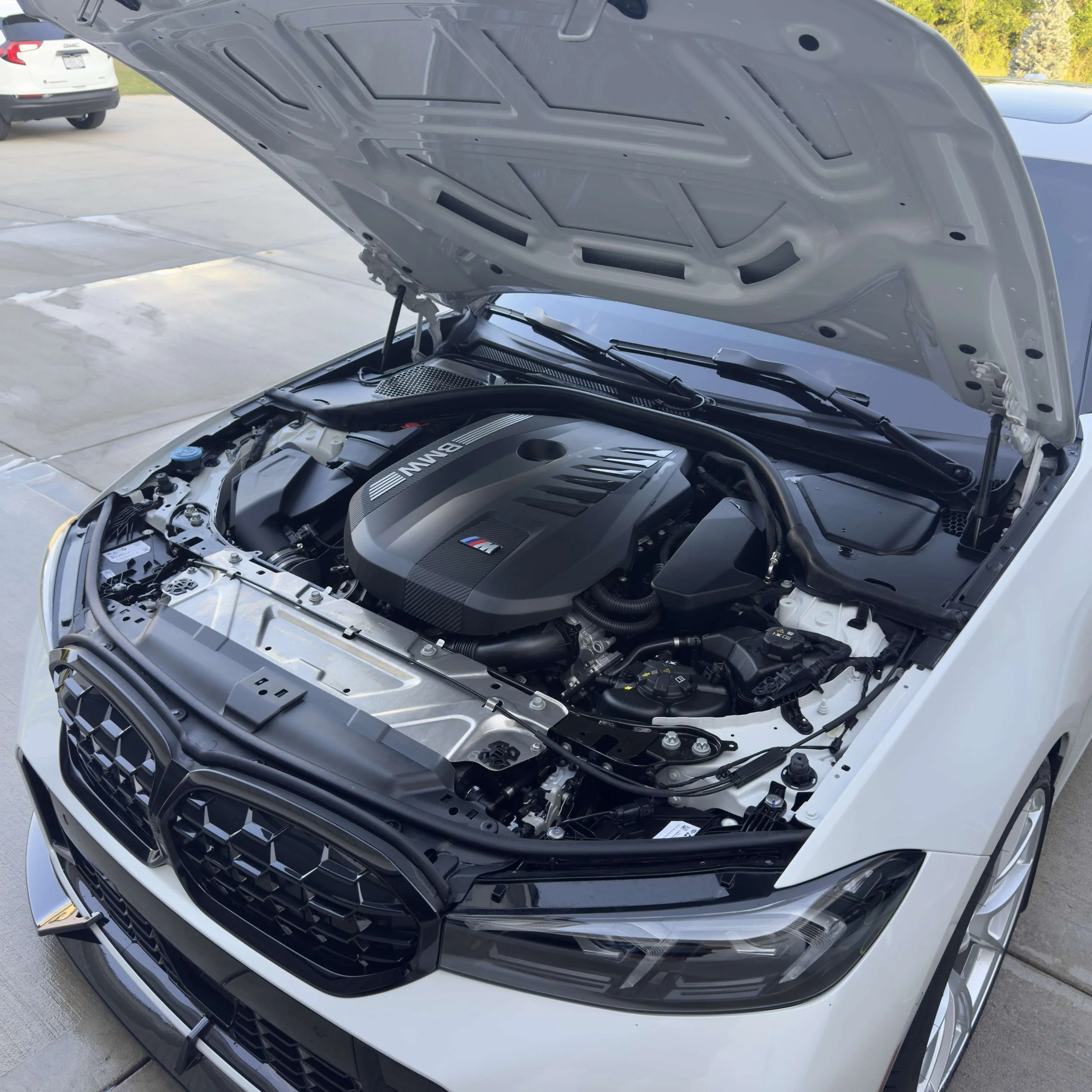 The engine of a white BMW car with its hood open, showing a clean, modern engine bay with BMW M branding visible on the engine cover.