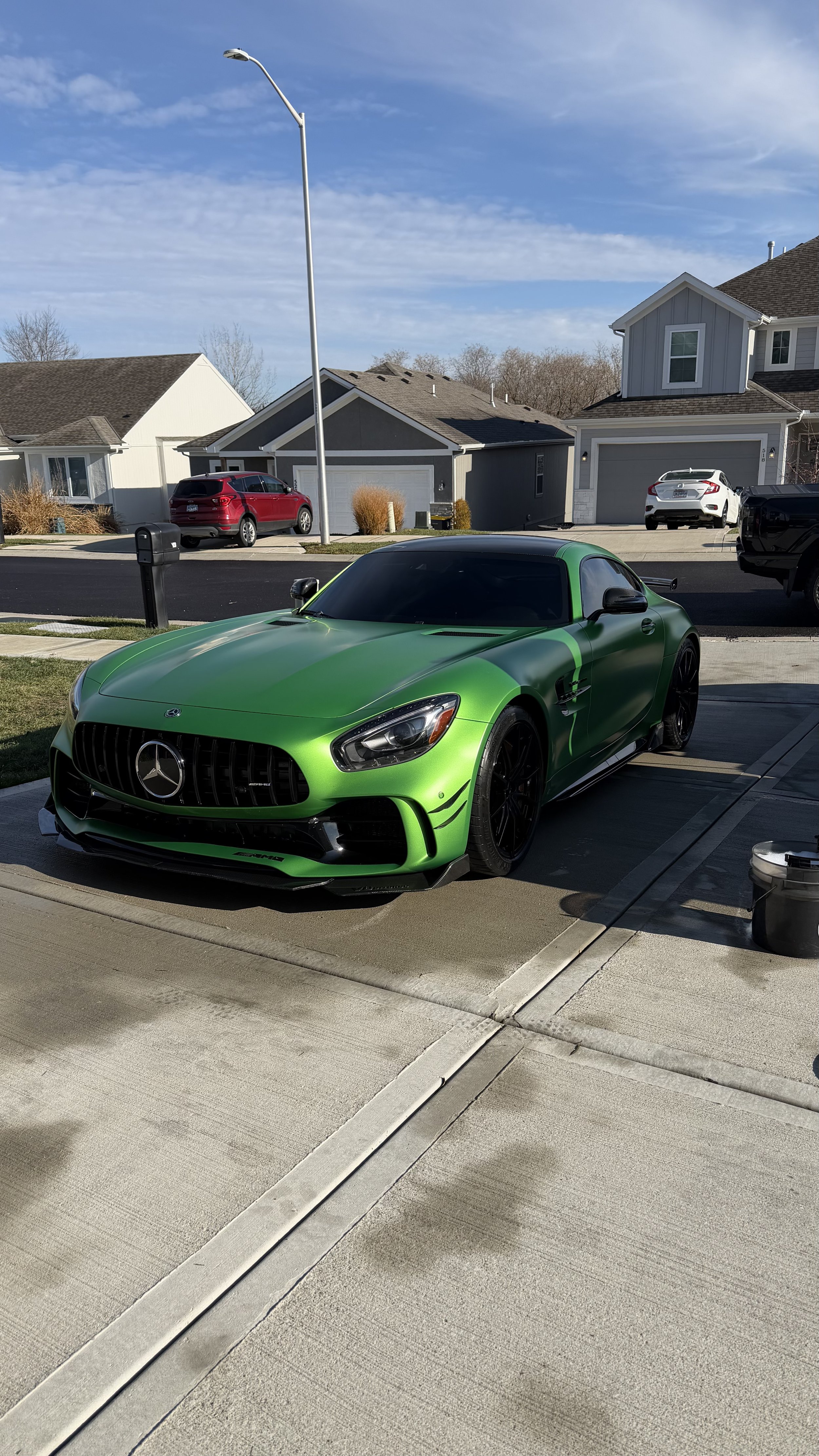 A green Mercedes-Benz sports car parked on a residential driveway, with houses and cars in the background during daytime.