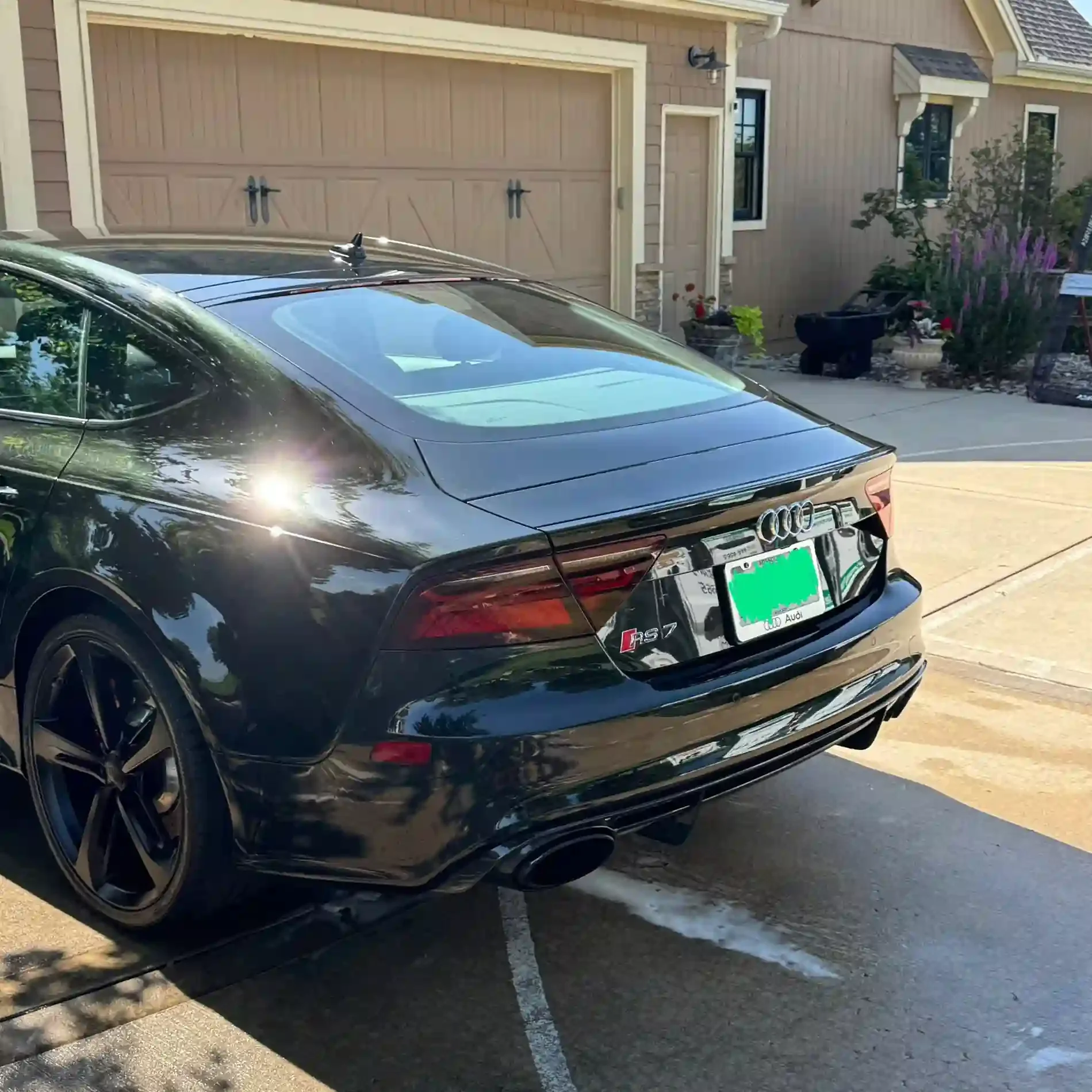 Black Audi RS7 parked in driveway outside a house with a tan garage door and garden with flowers.