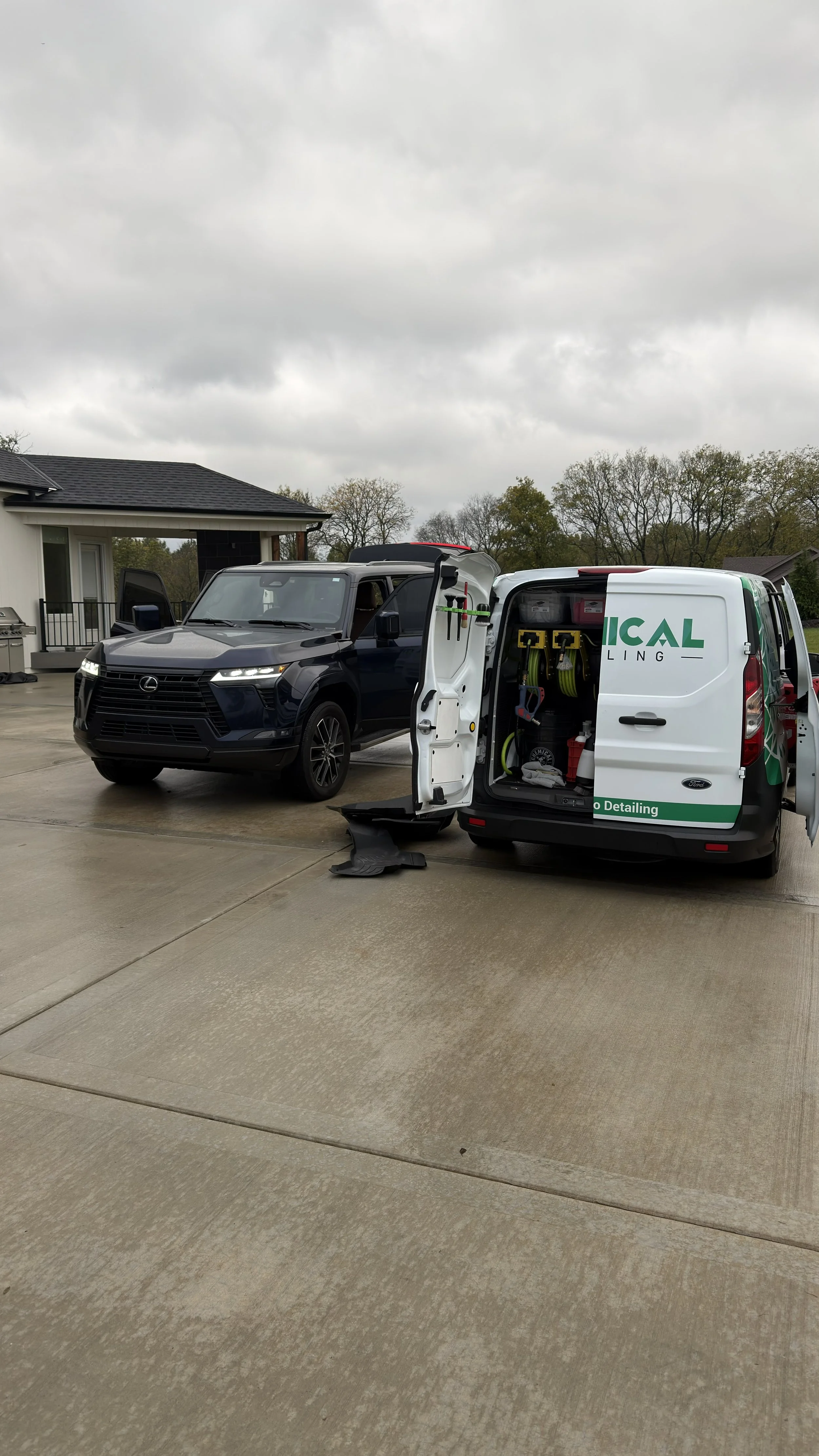 A black Lexus SUV parked on a driveway next to a white service van with its side door open, revealing tools and equipment inside. The driveway is wet under a cloudy sky.