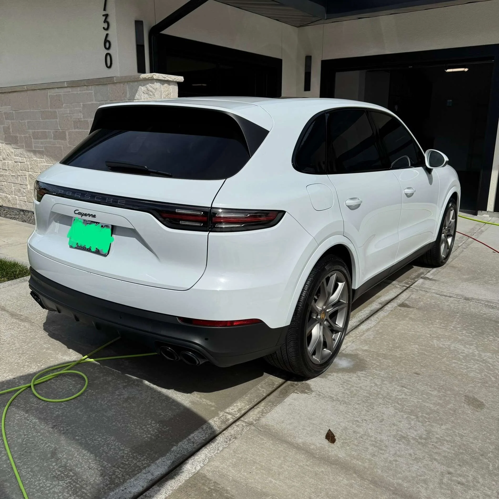 White Porsche SUV parked outside a modern house, connected to a green garden hose.