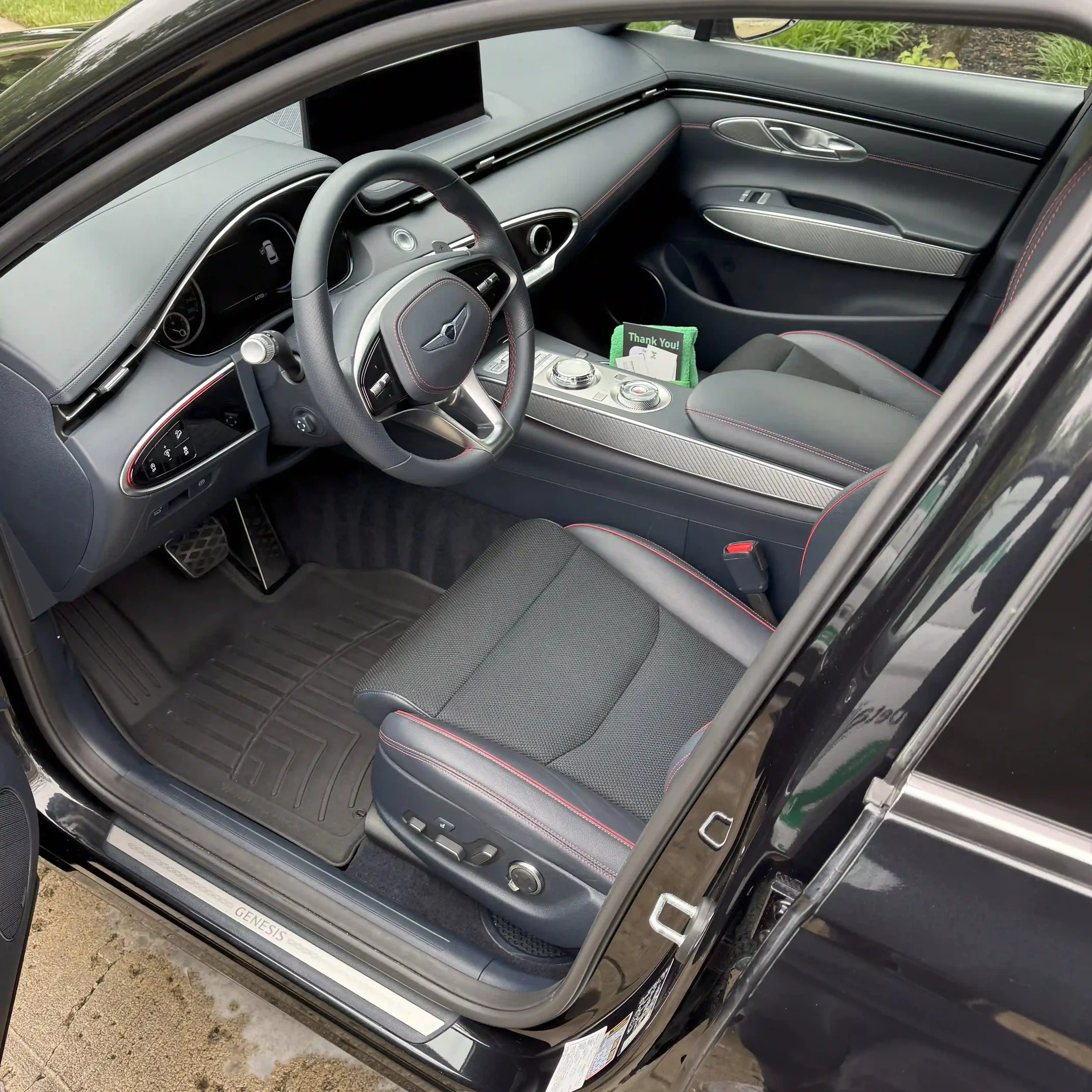Interior of a black Genesis car showing the driver's seat, steering wheel, center console with control buttons, and a thank you card in the door pocket.