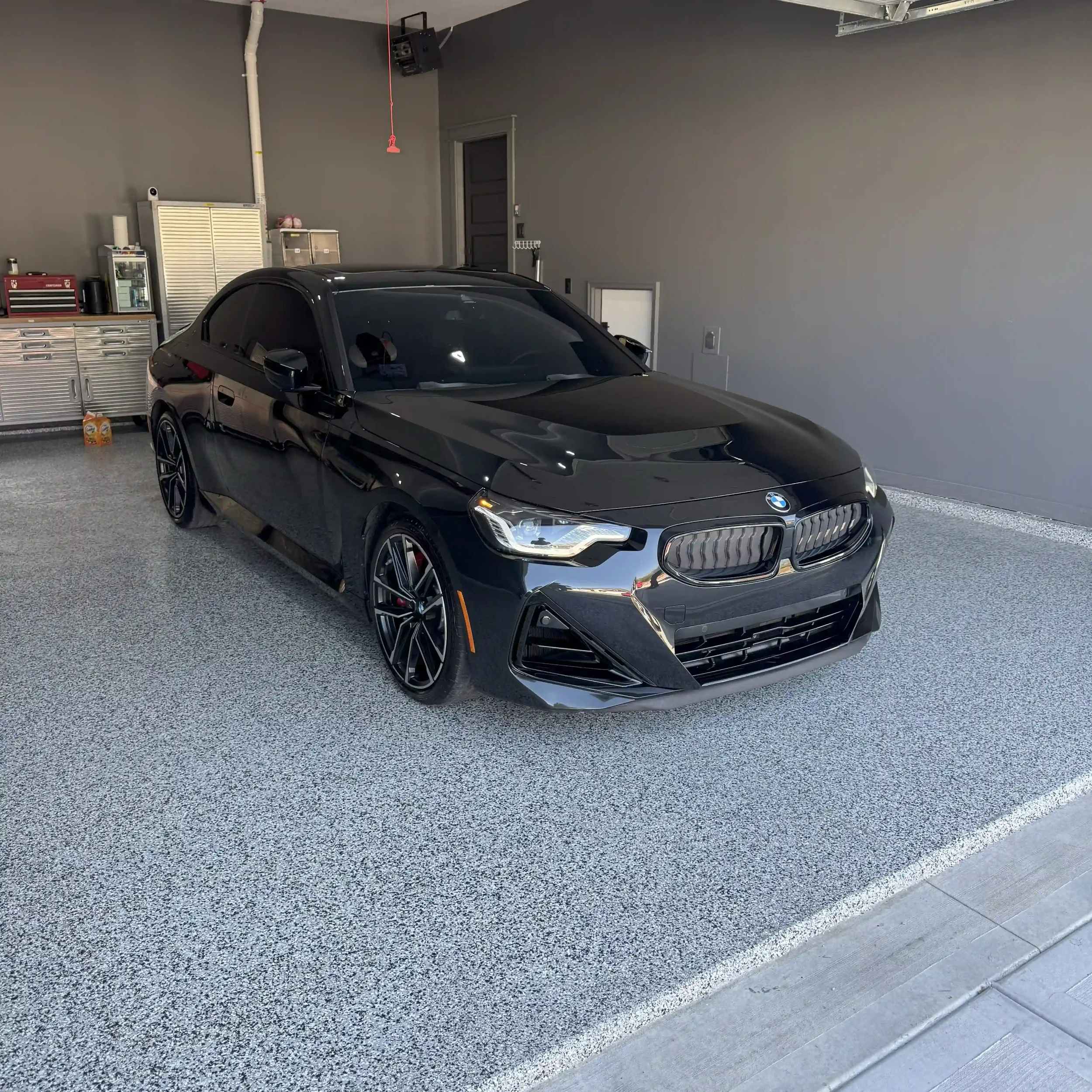 Black sports car parked in a clean garage with gray walls and storage cabinets in the background.