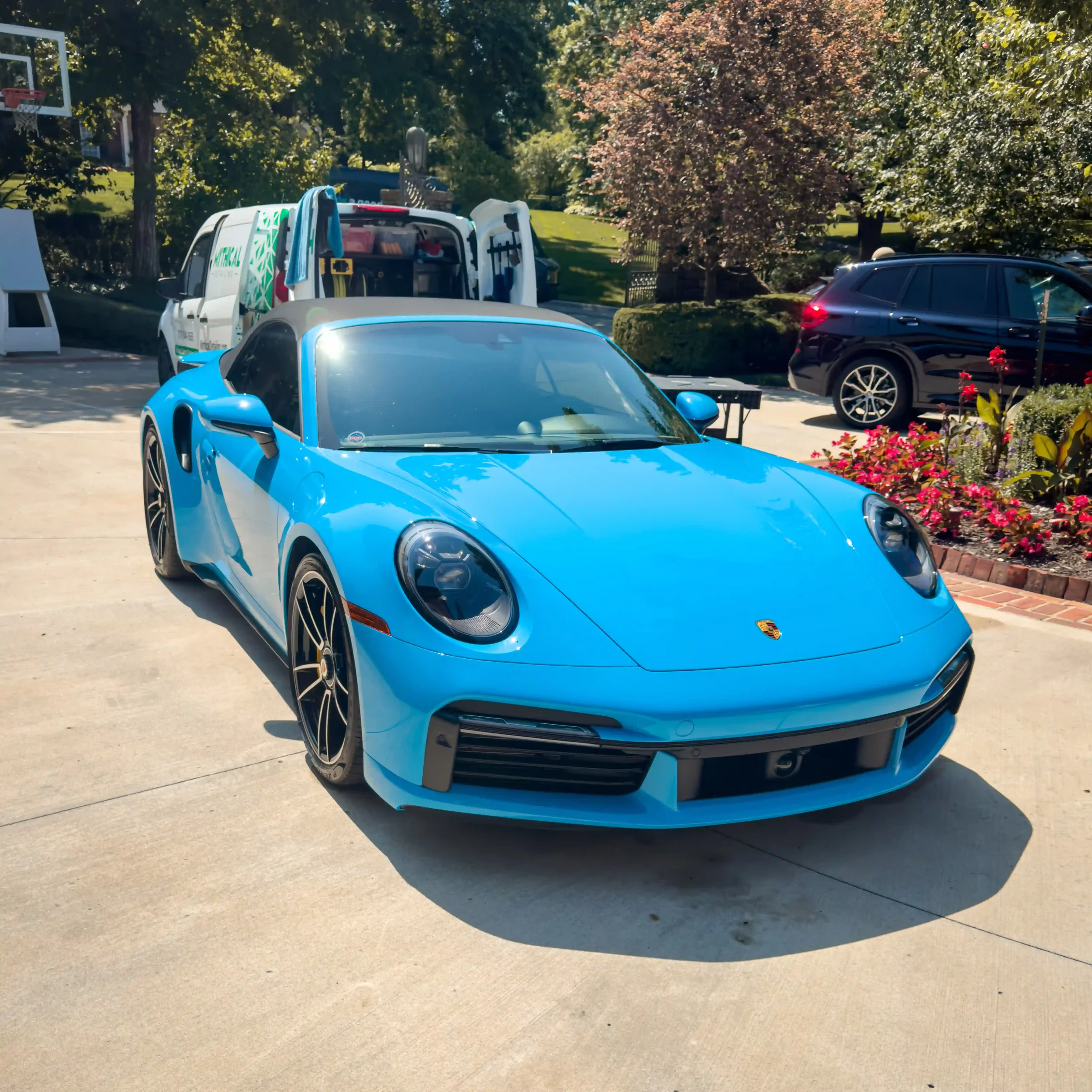 Blue Porsche sports car parked on driveway with flowering bushes and trees in background.