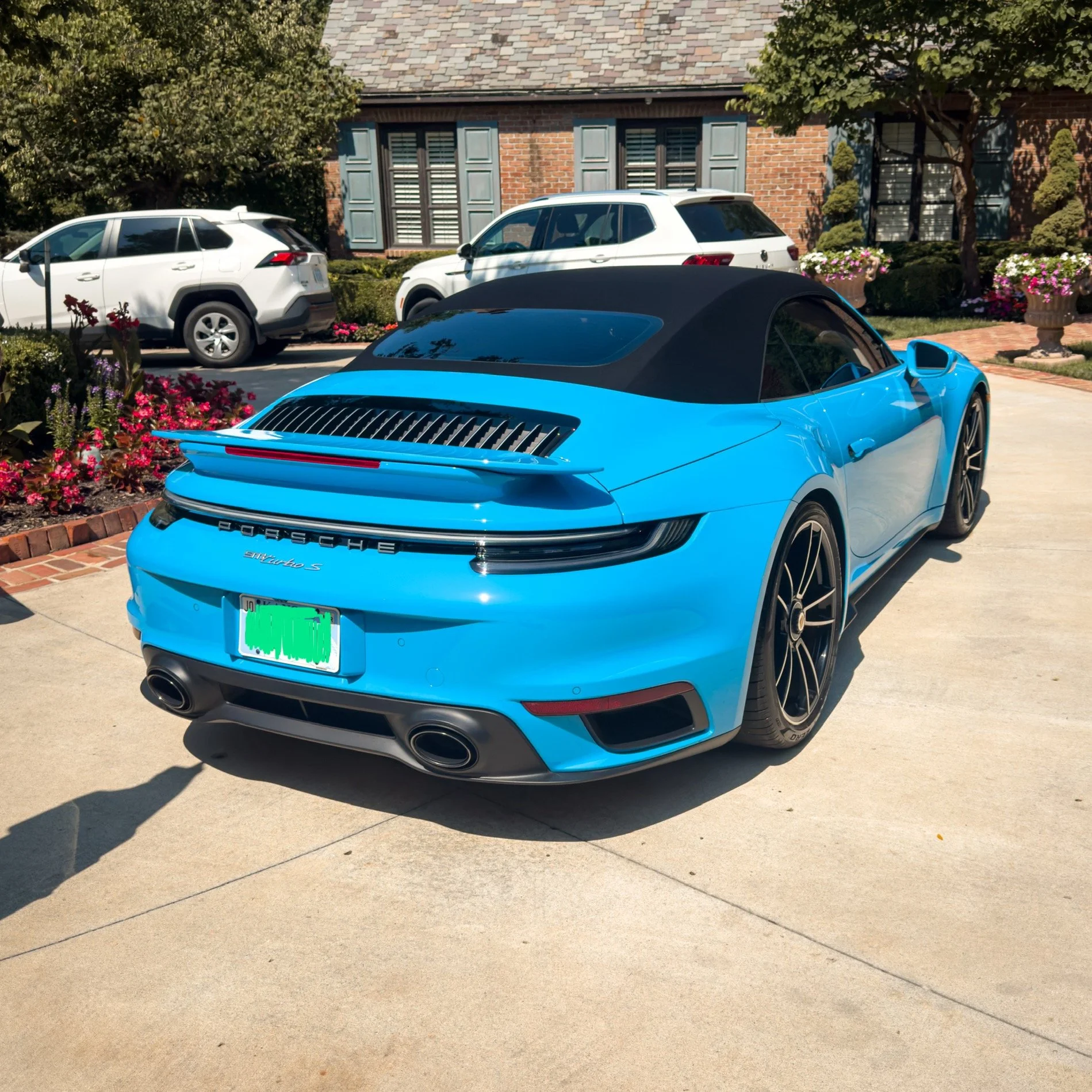 Blue Porsche 911 convertible parked on driveway with other cars and a house in the background.