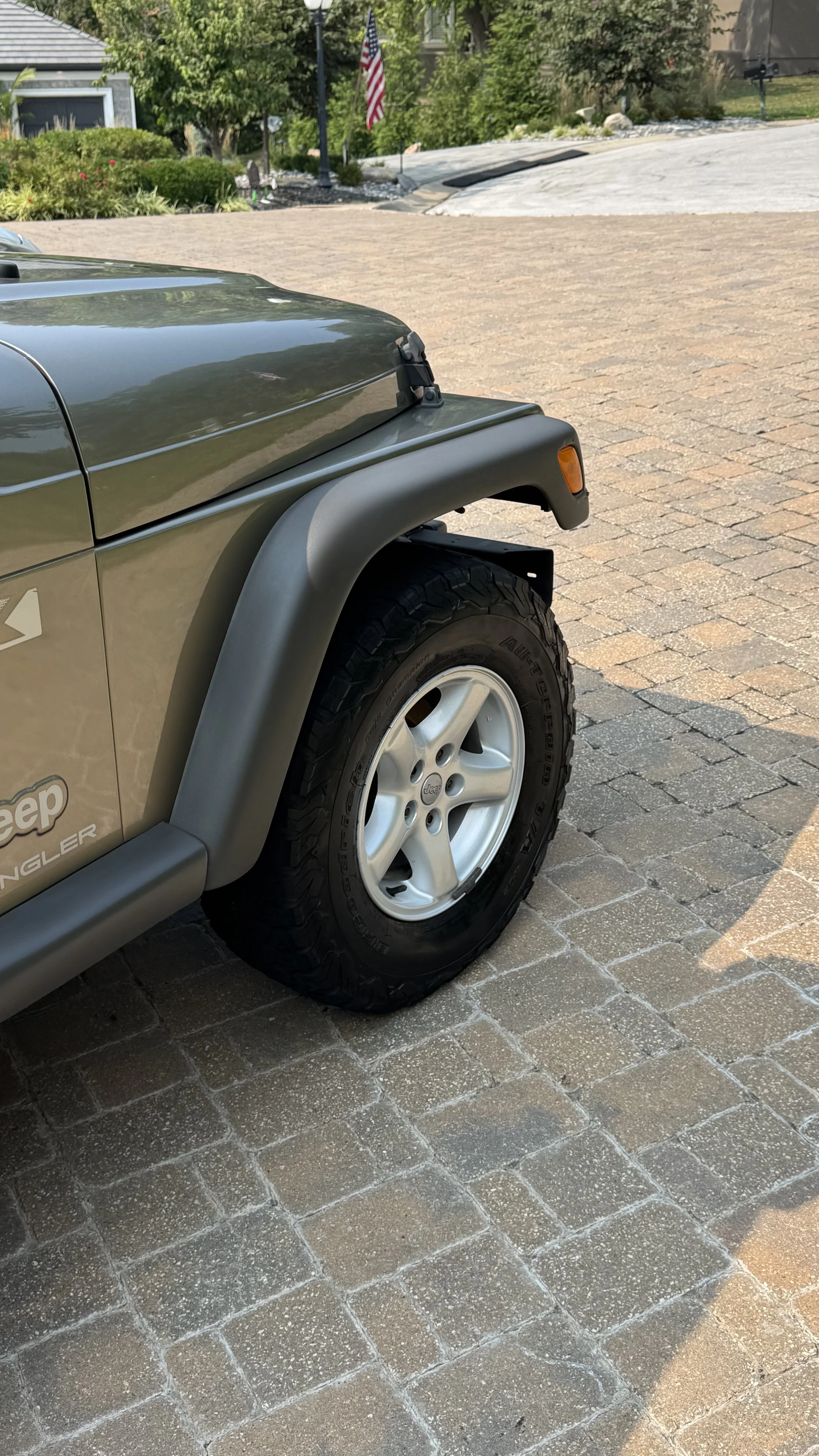 A green Jeep parked on a brick driveway with a garden, trees, and an American flag in the background.