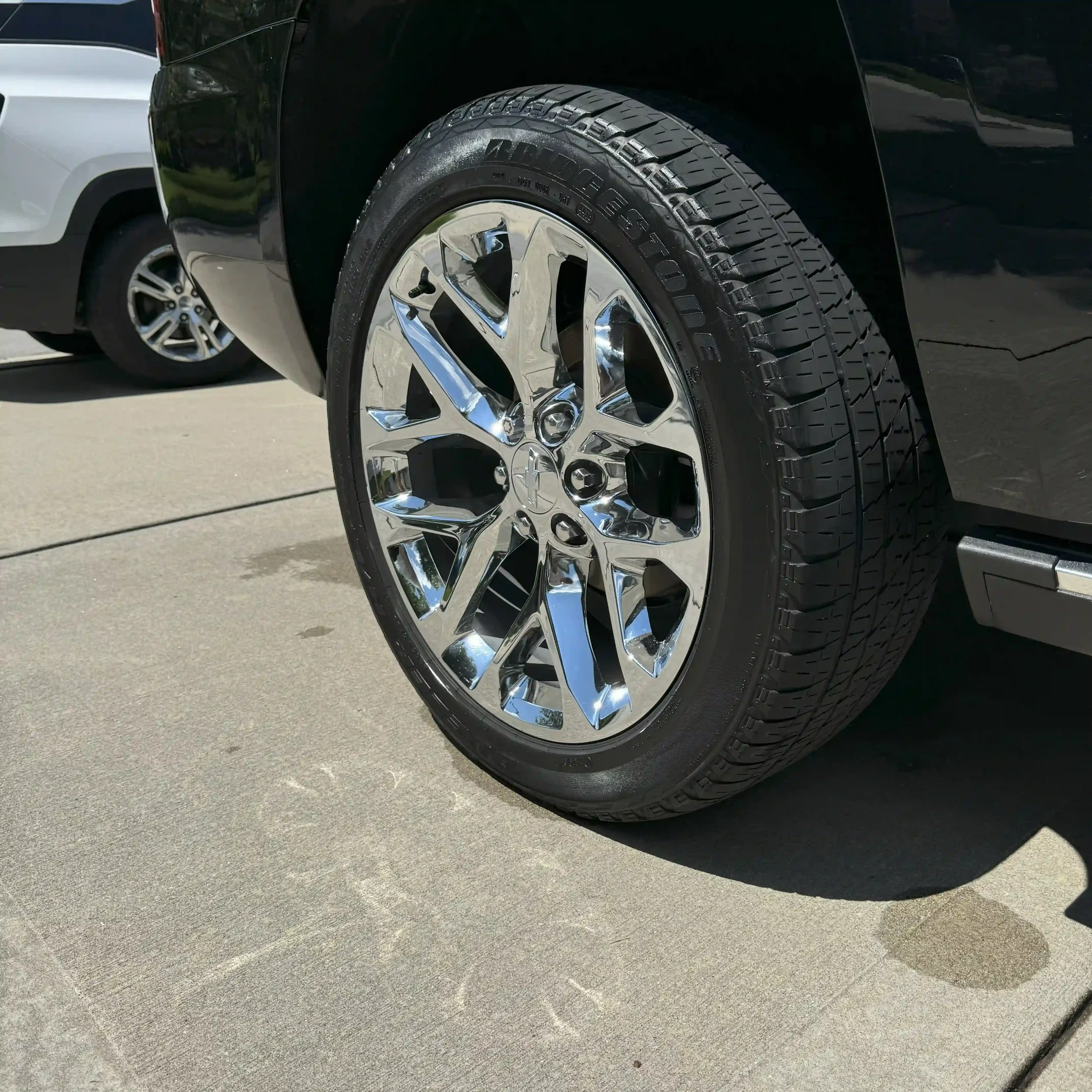 Close-up of a shiny chrome wheel with an all-terrain tire on a black vehicle parked on a concrete driveway.