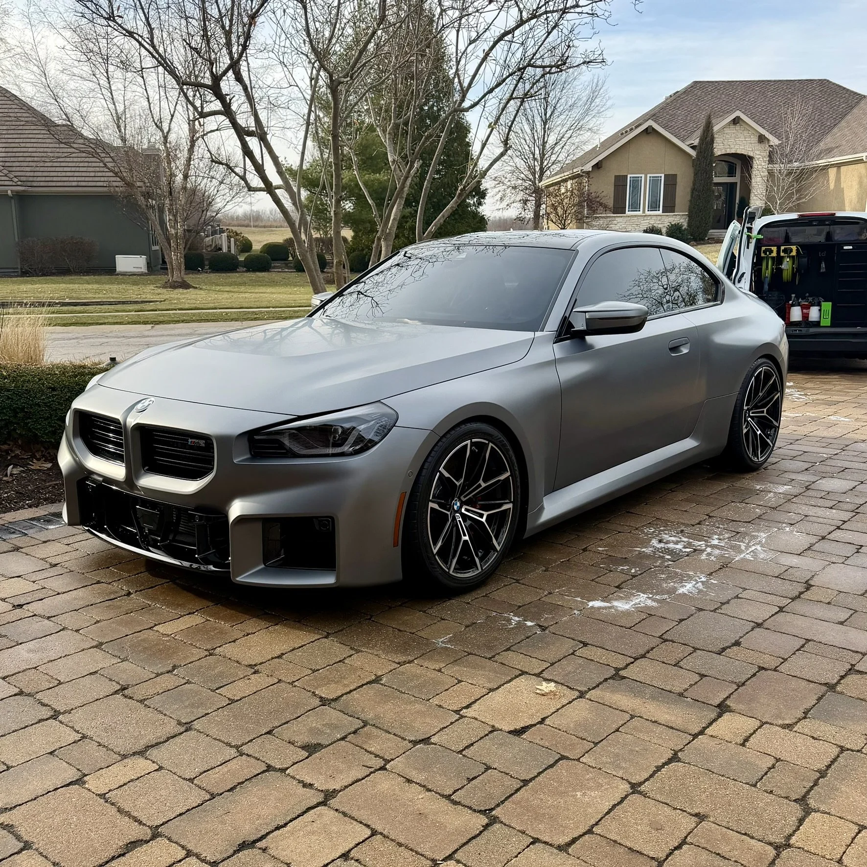 A silver BMW M4 parked on a brick driveway in front of a suburban house with leafless trees, and a vehicle with open doors in the background.