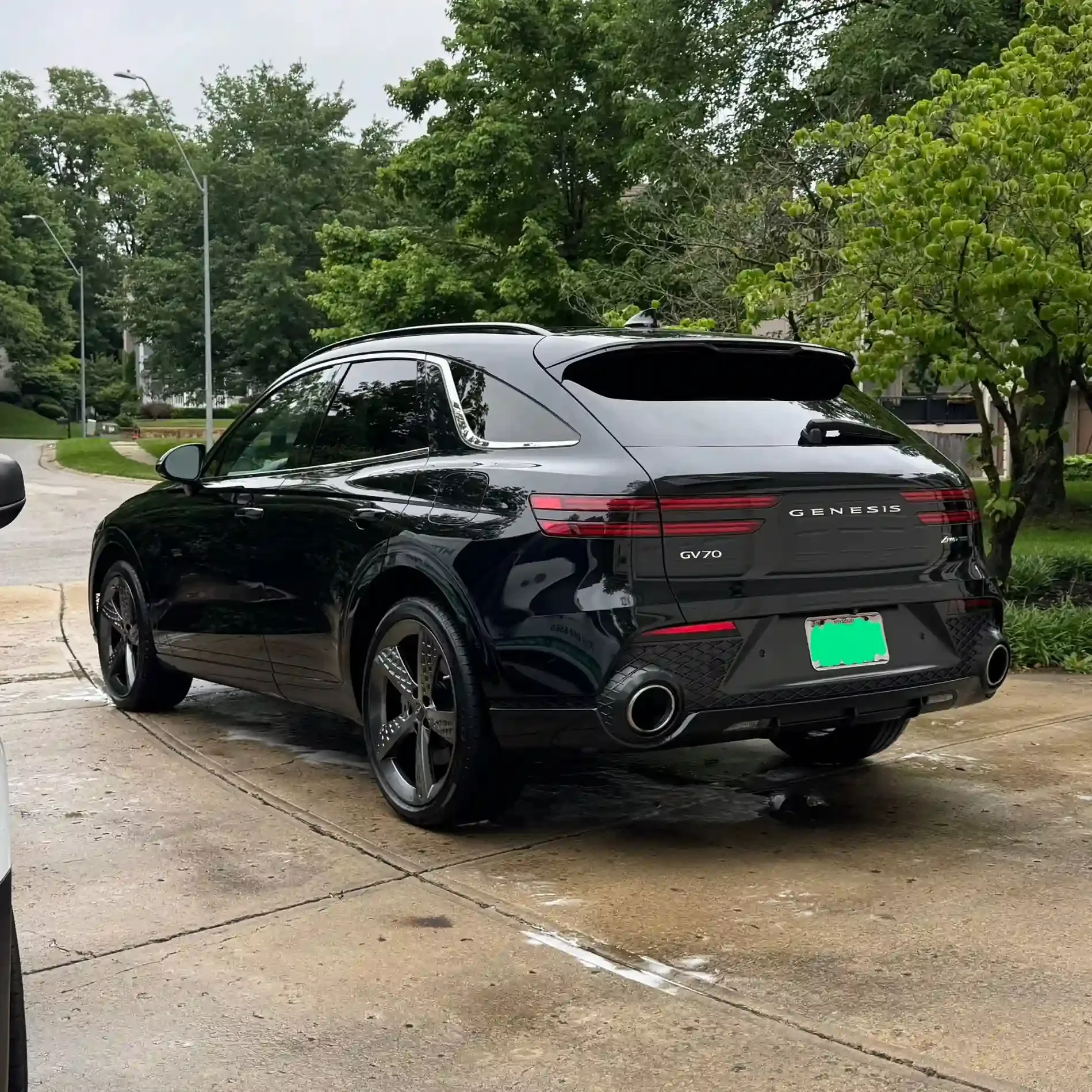 Black Genesis GV70 luxury SUV parked on a wet driveway, surrounded by green trees and grass.