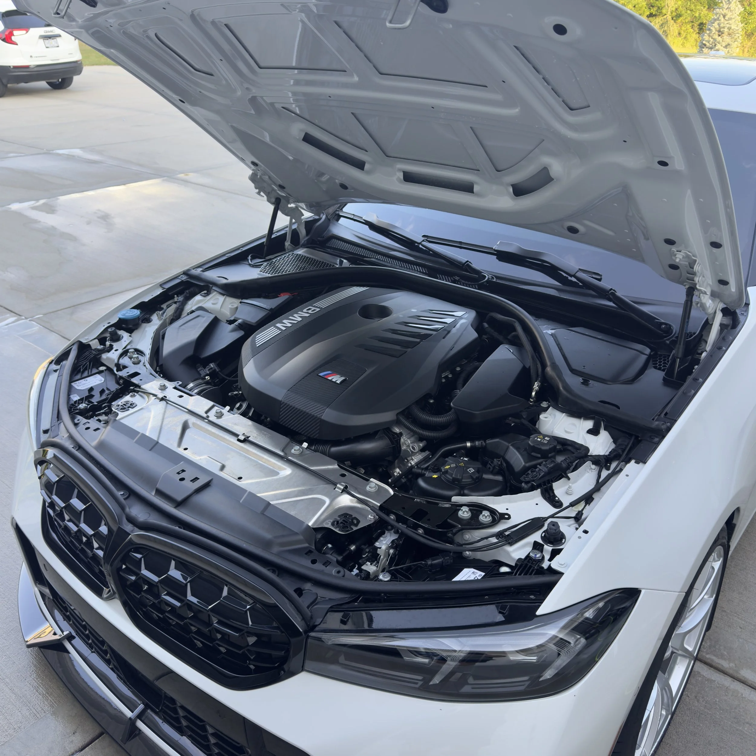 White car with open hood revealing BMW engine with M badge in a driveway.