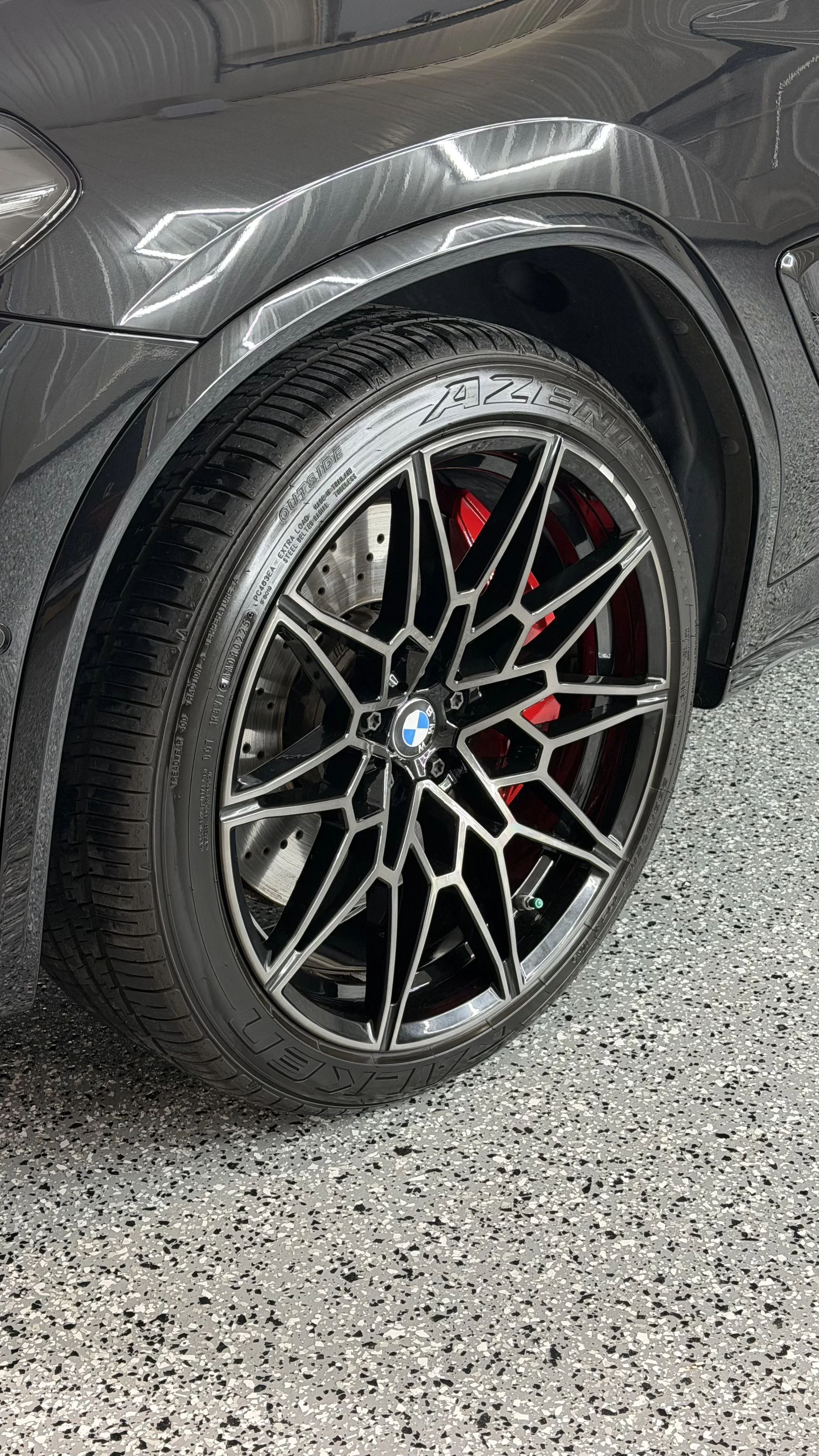 Close-up of a BMW vehicle wheel with a black and silver intricate rim, red brake caliper, and tire branded Falken AZENIS on a speckled gray garage floor.