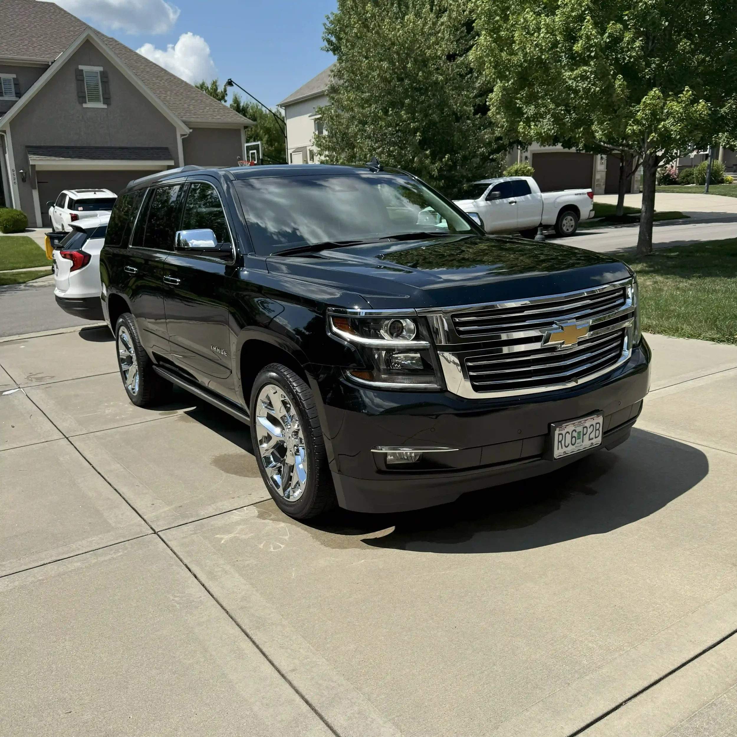 Black Chevrolet Tahoe SUV parked on a driveway in a suburban neighborhood with other vehicles, trees, and houses in the background.