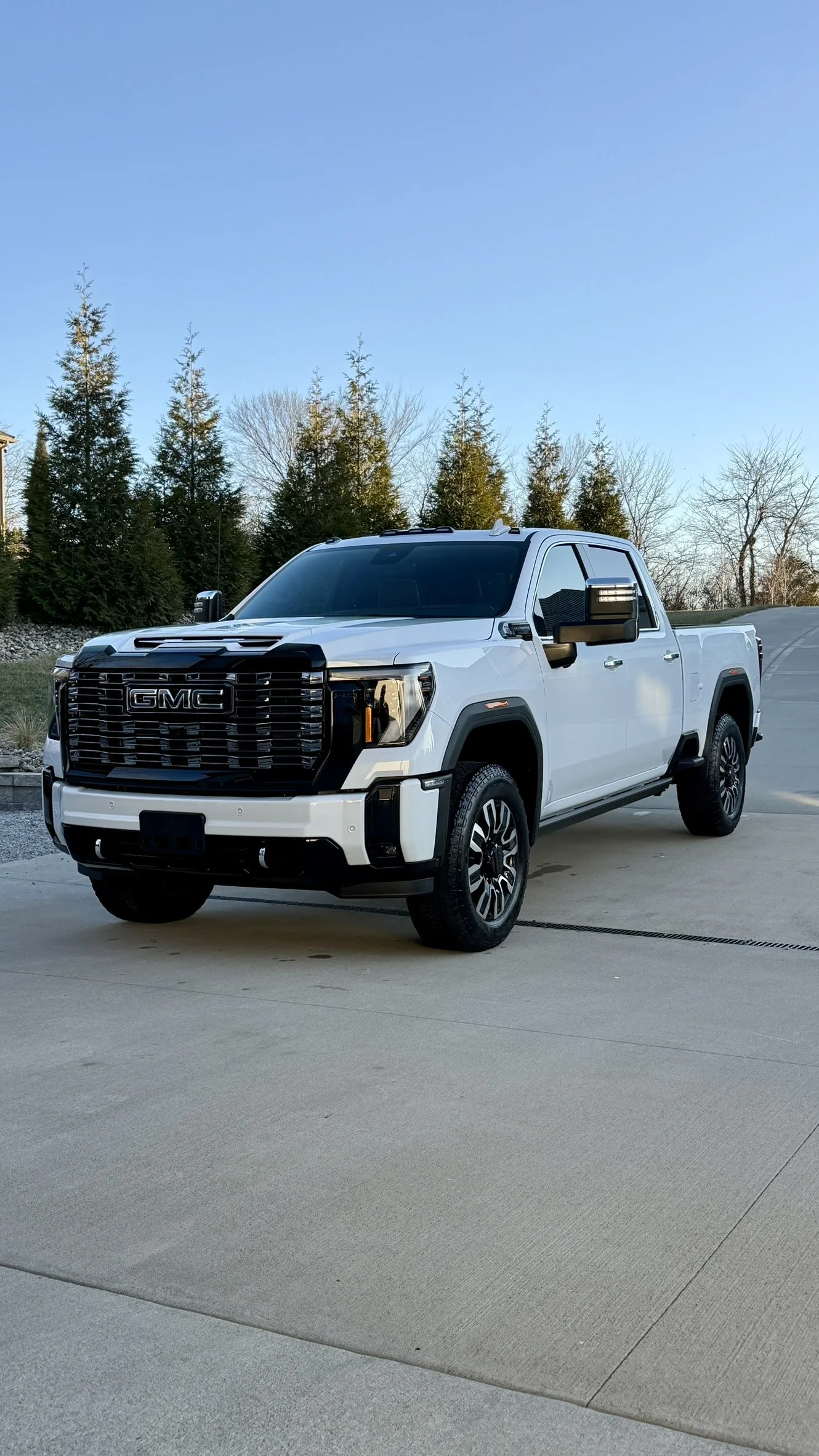A white GMC pickup truck parked on a concrete driveway with trees and a clear blue sky in the background.