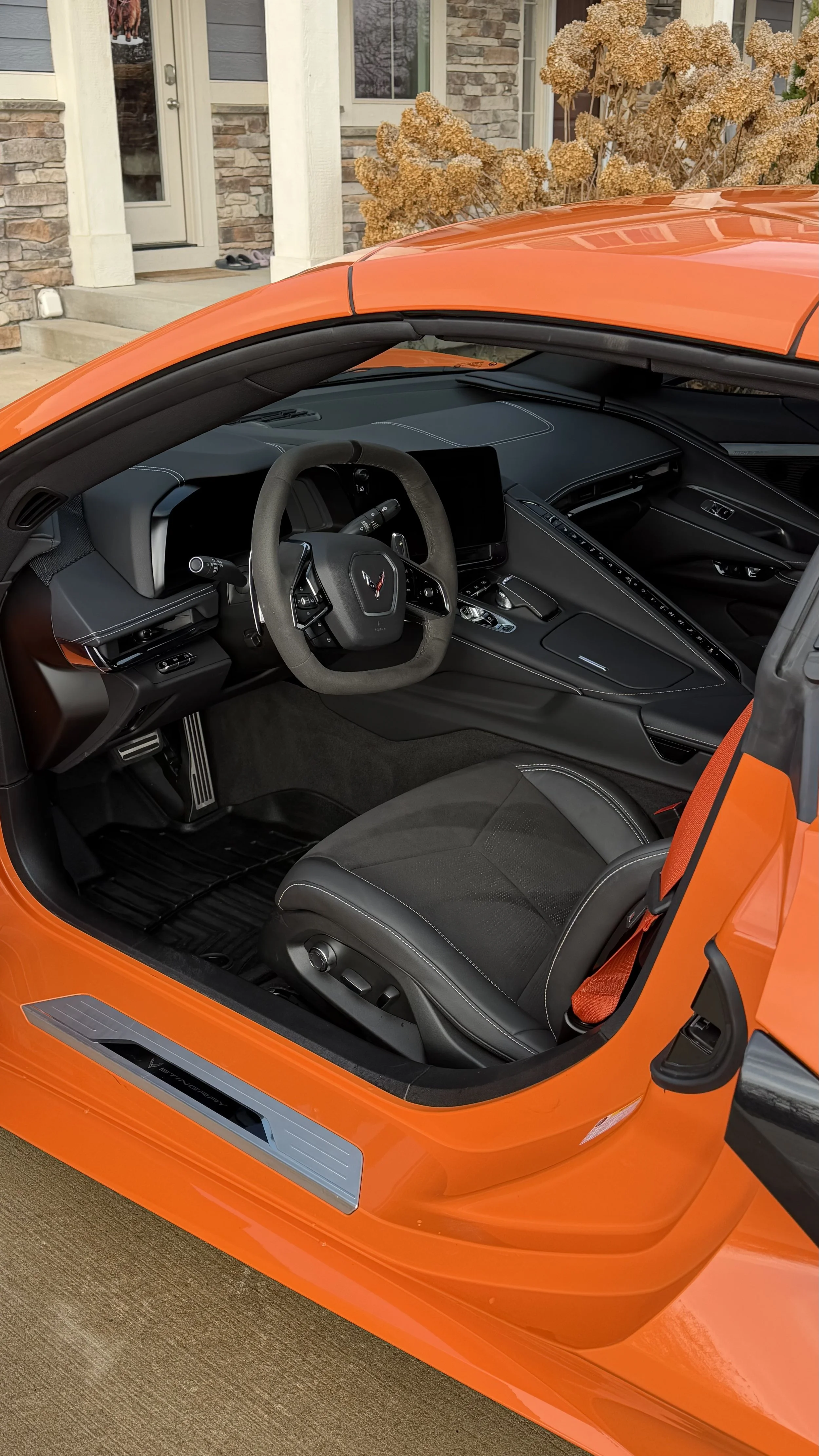 Interior of an orange sports car with black seats, steering wheel, dashboard, and a digital display, parked outside a house with flowers in the background.