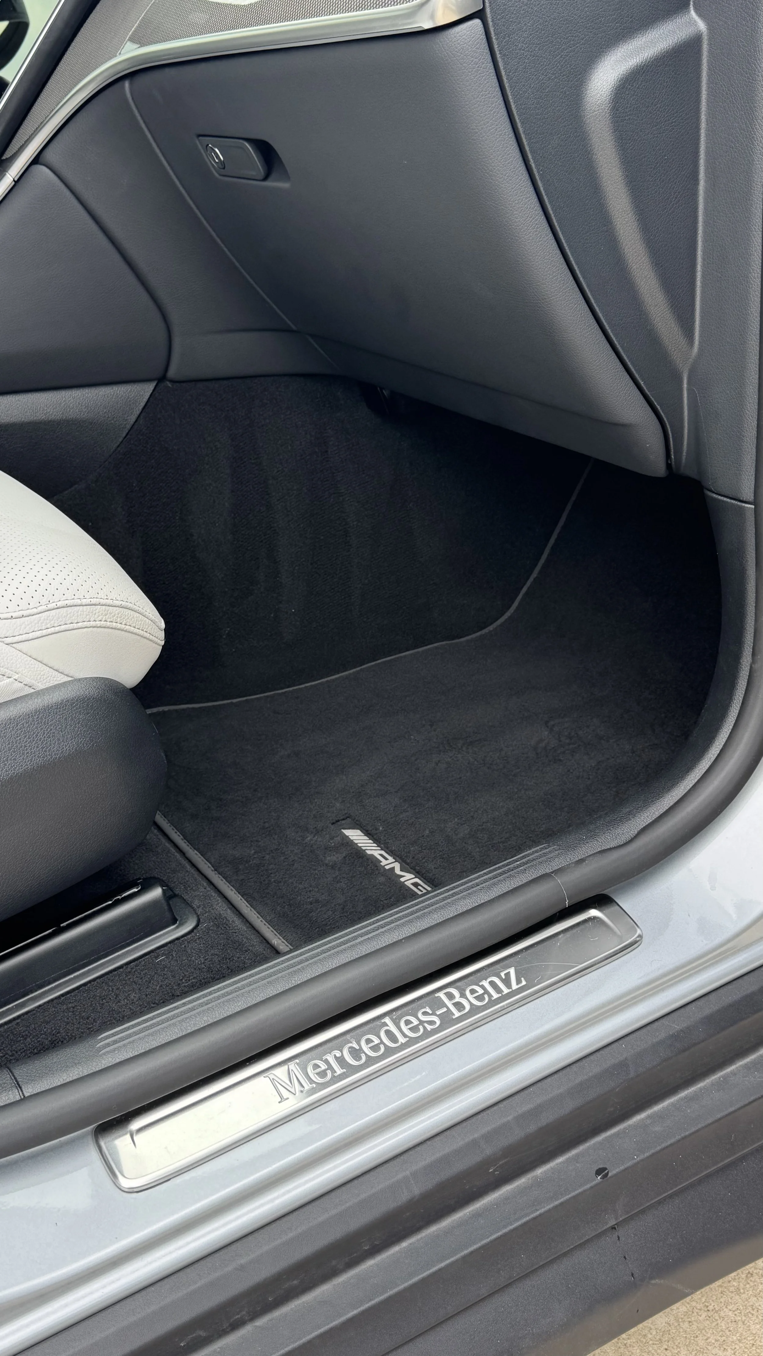 Interior of a car showing the passenger footwell with a black carpet mat that has an AMG logo. The door sill has a metallic plate with the Mercedes-Benz logo and branding.
