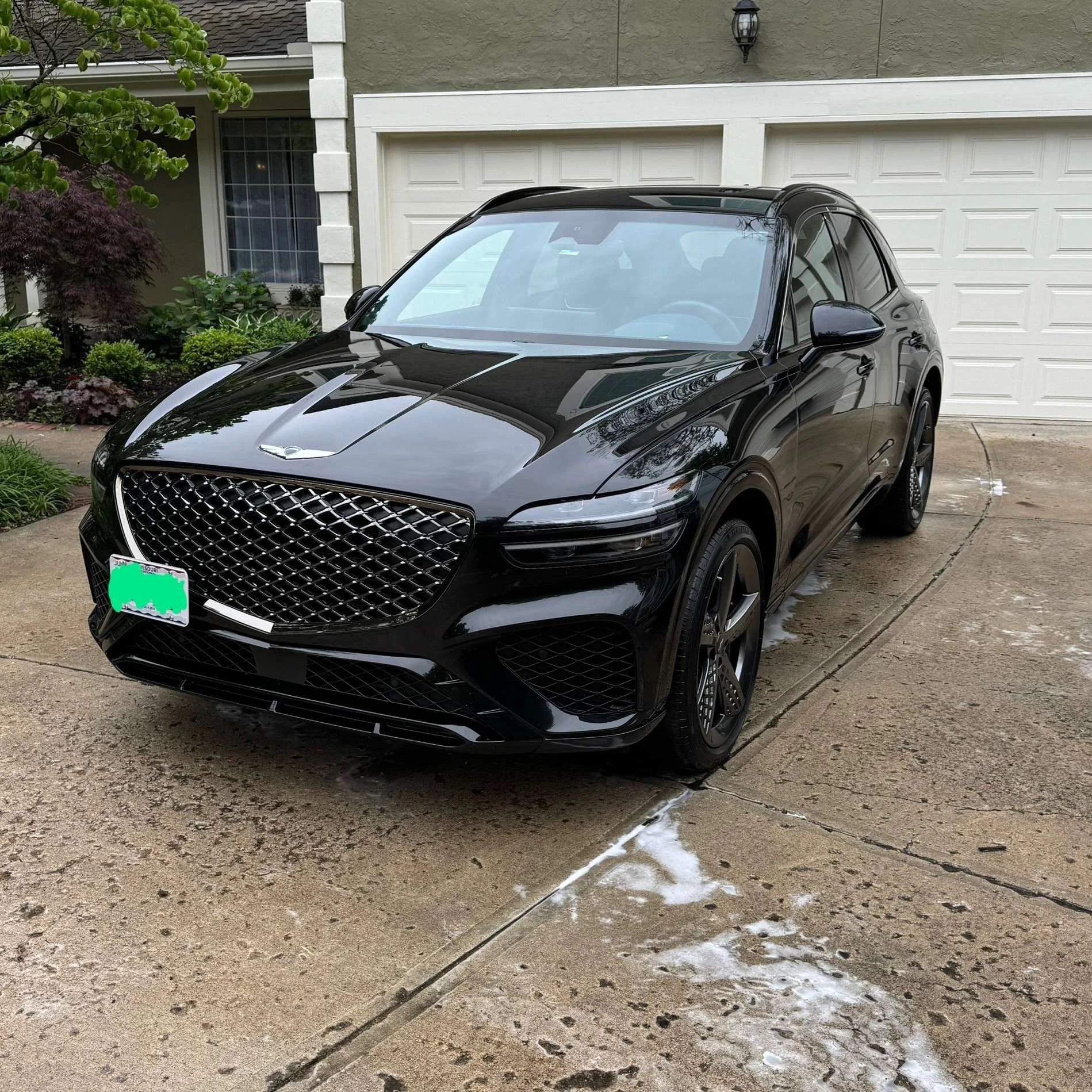 Black luxury SUV parked on a concrete driveway near a white garage door and a house with plants and greenery nearby.