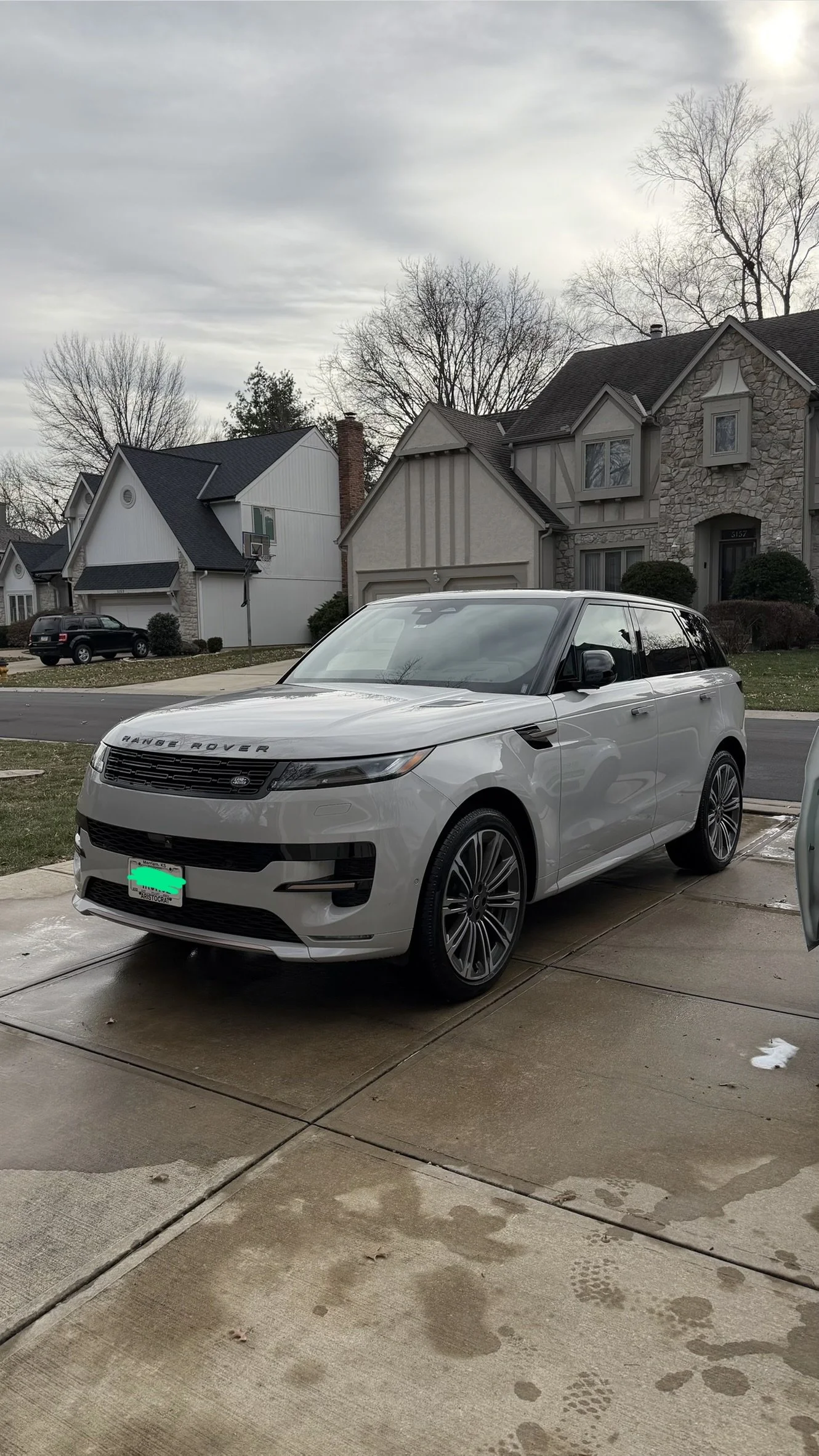 A white Range Rover parked on a driveway in front of a residential neighborhood with houses and leafless trees under a cloudy sky.