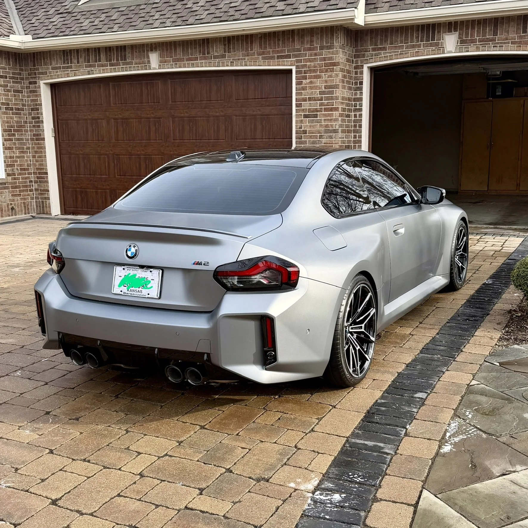 A silver BMW M2 sports car parked on a brick driveway outside a house with a brick exterior and a wooden garage door.