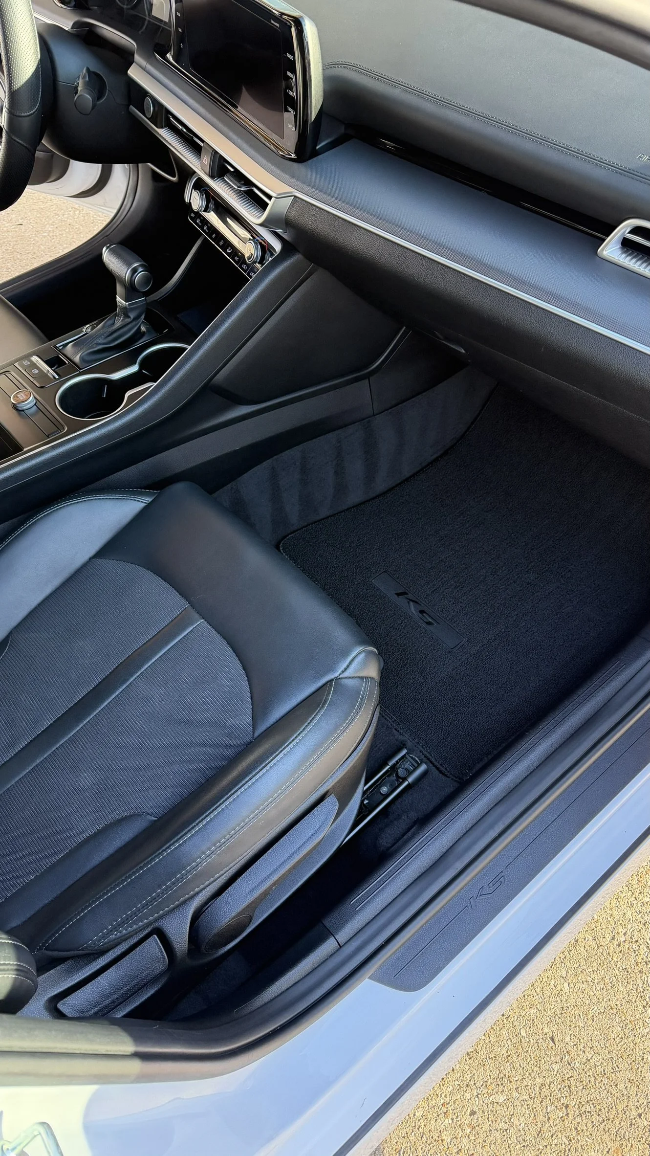 Interior of a modern car showing the front passenger seat, dashboard with a touchscreen display, gear shifter, cup holders, and a floor mat with the car's logo.