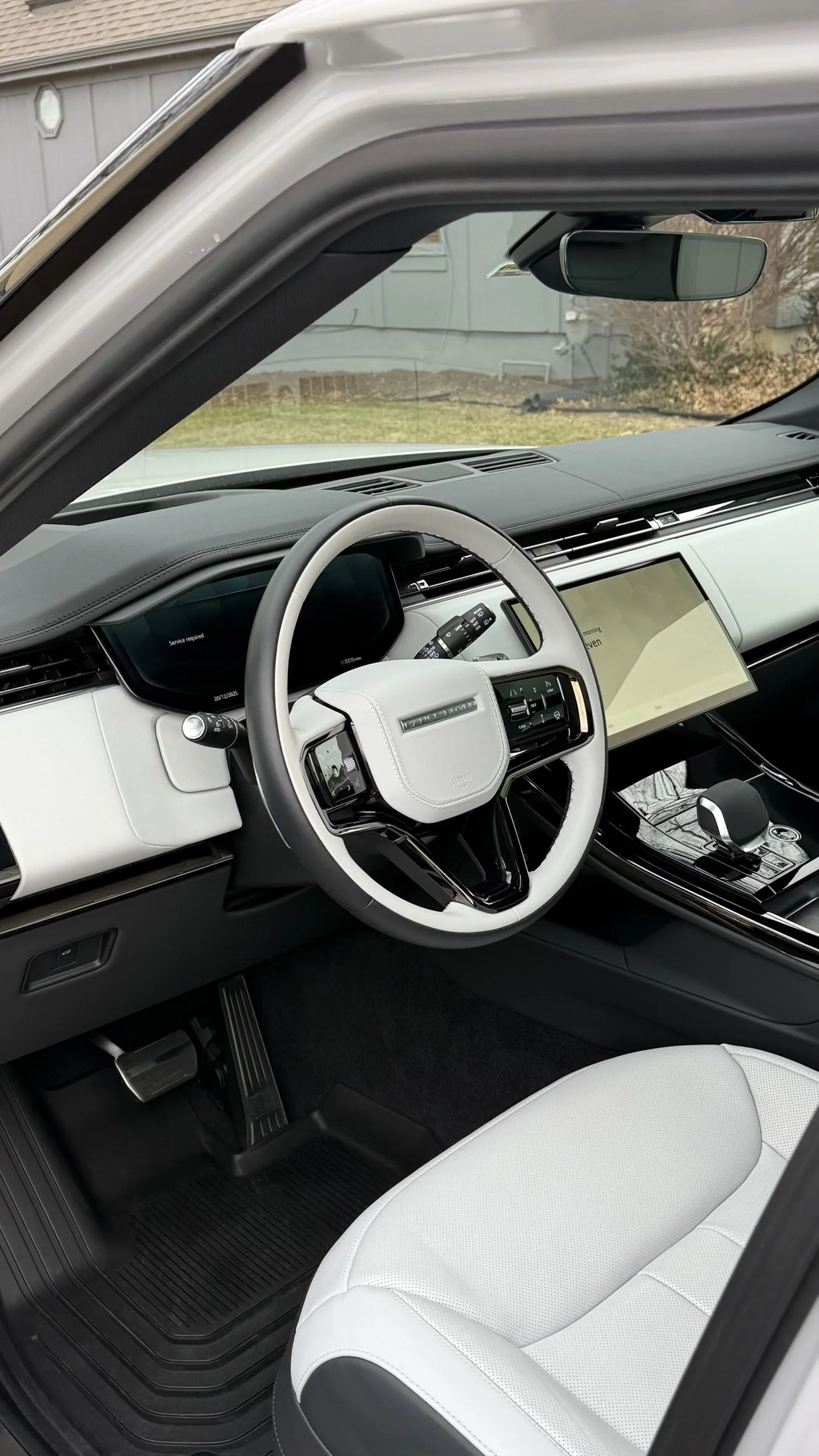 Interior of a modern vehicle showing a white and black dashboard, a touchscreen display, and a white and black steering wheel. The vehicle has a light-colored seat and a view of the front windshield with a house and yard outside.