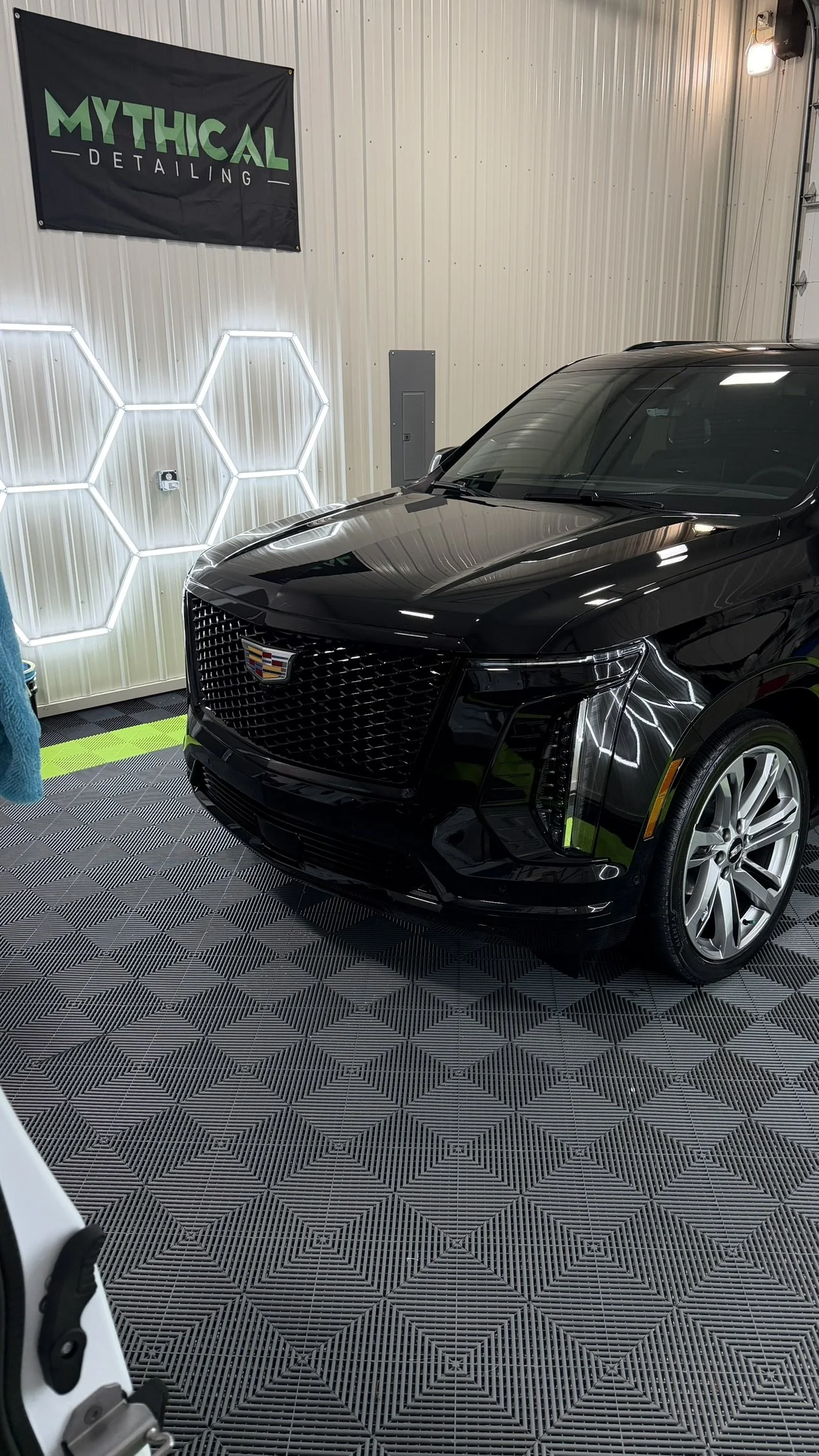 Black Cadillac SUV inside a detailing garage with illuminated hexagon wall decor and a 'Mythical Detailing' banner on the wall.