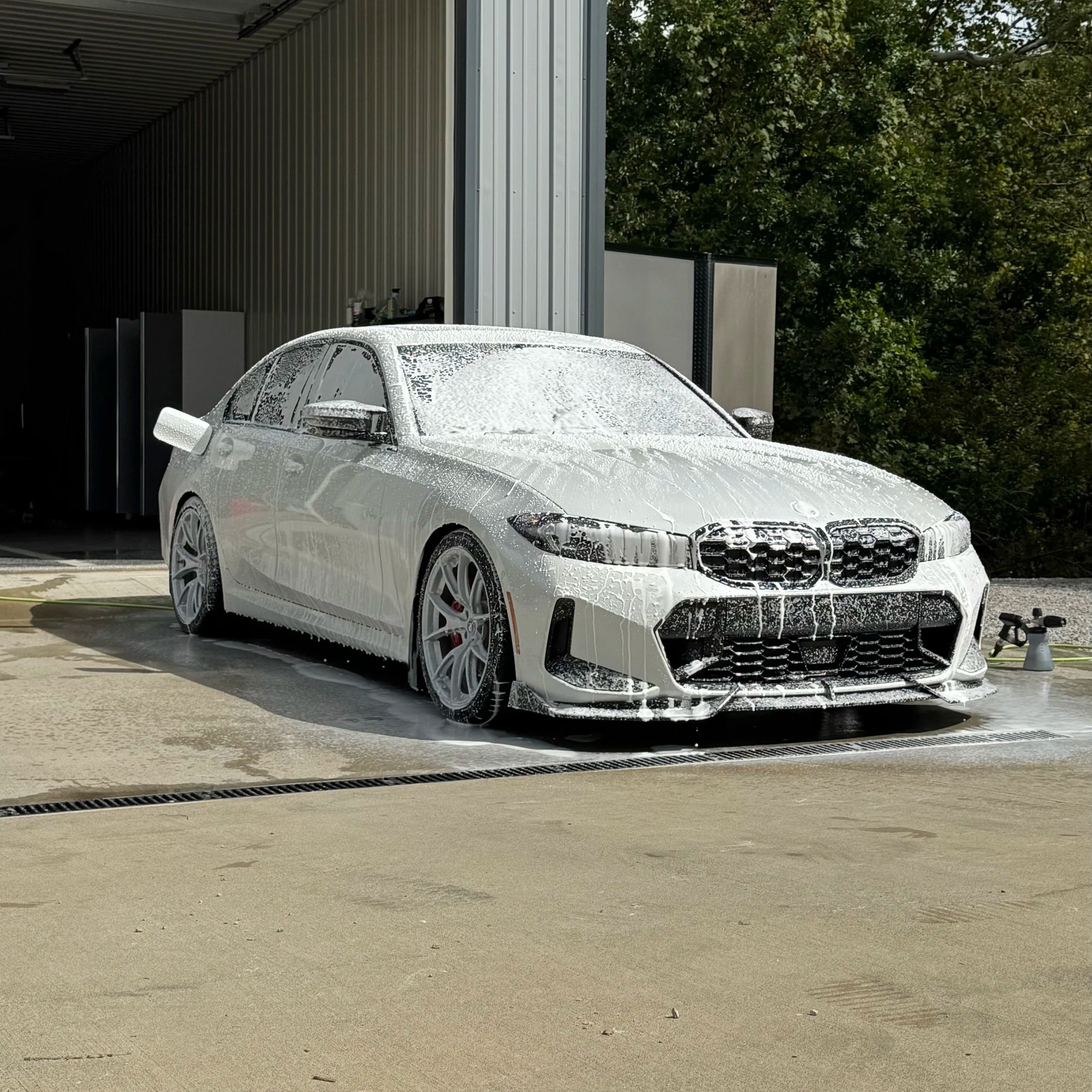 A white sports sedan being washed with soap and foam, outside near a metal building and trees in the background.