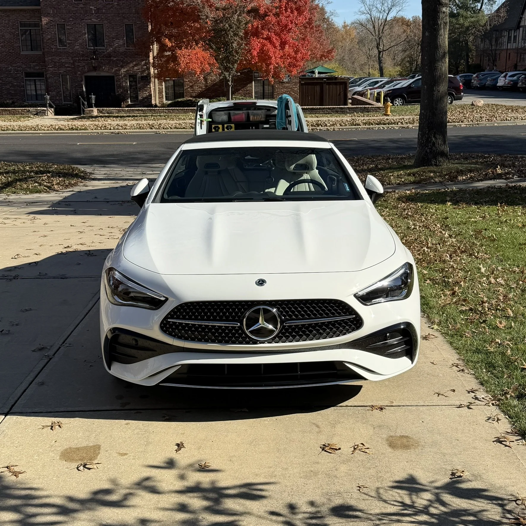 Front view of a white Mercedes-Benz car parked on a sidewalk with trees and residential buildings in the background.