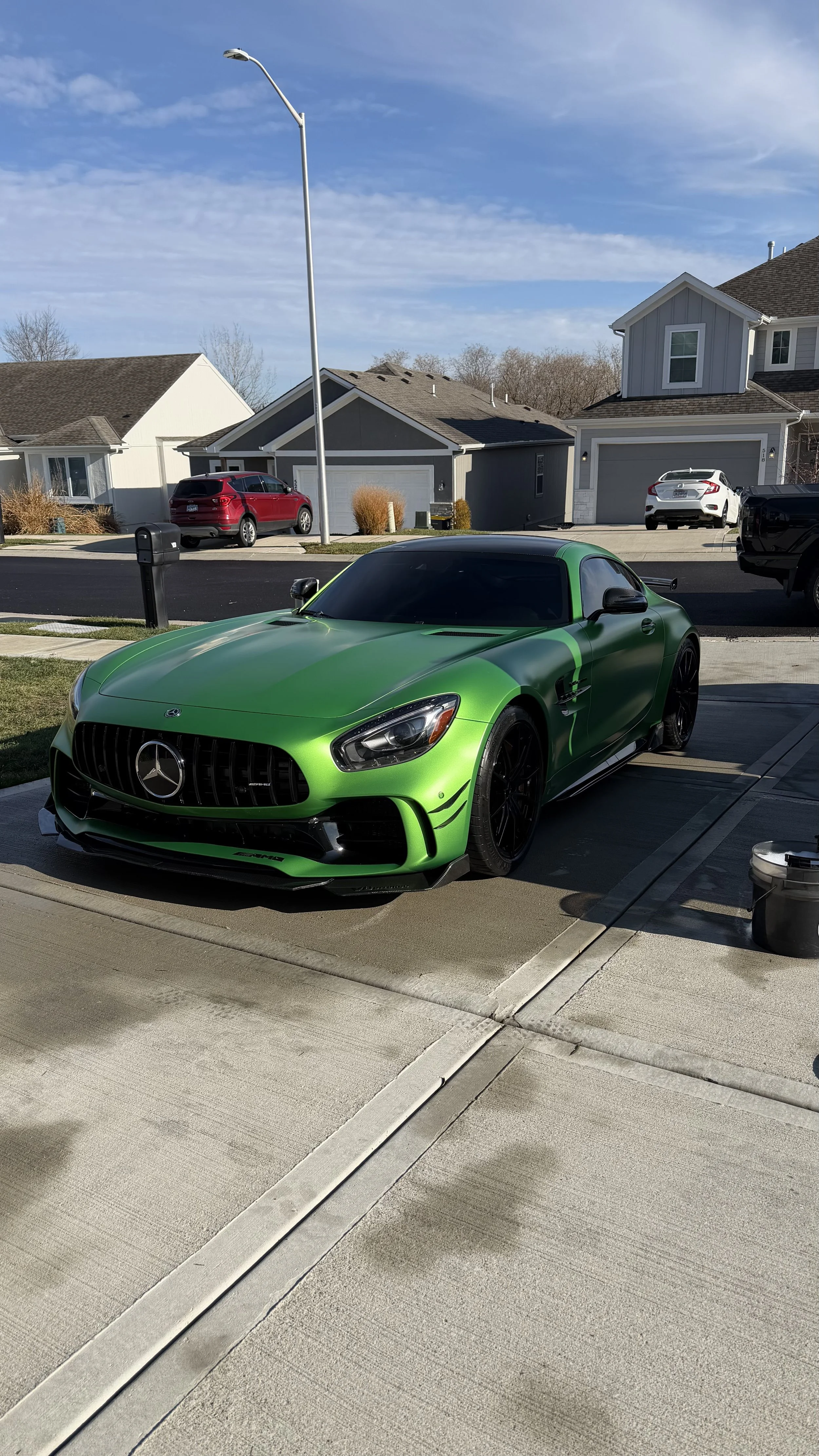 Green Mercedes-Benz sports car parked on driveway in front of suburban house, with other cars and houses in background.