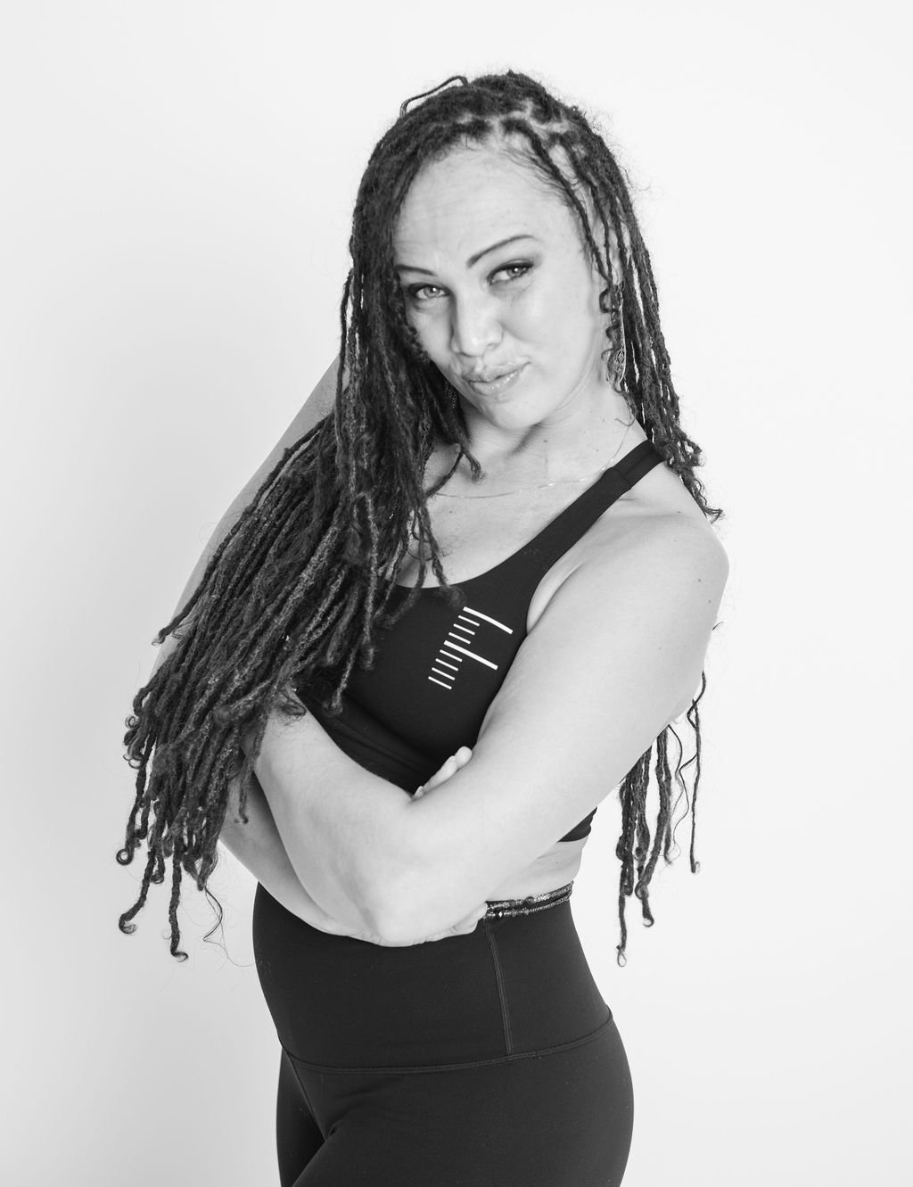Black and white photo of a woman with long dreadlocks, crossing her arms and smiling slightly, wearing a sports bra and leggings, standing against a plain white background.