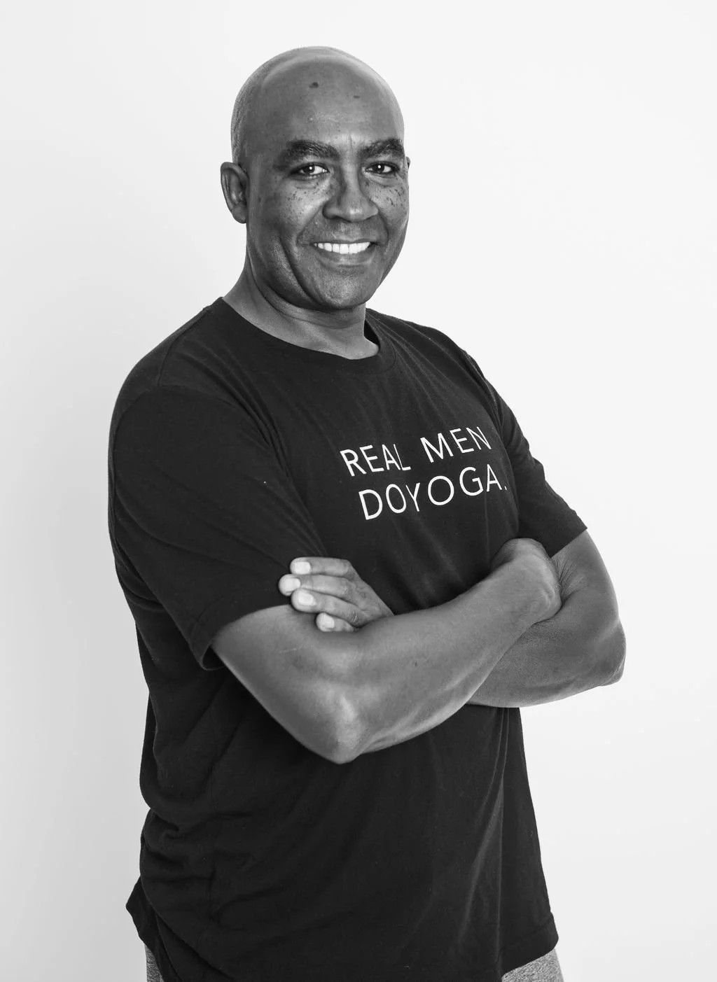 A smiling man with crossed arms wearing a black T-shirt that says 'REAL MEN DO YOGA'.