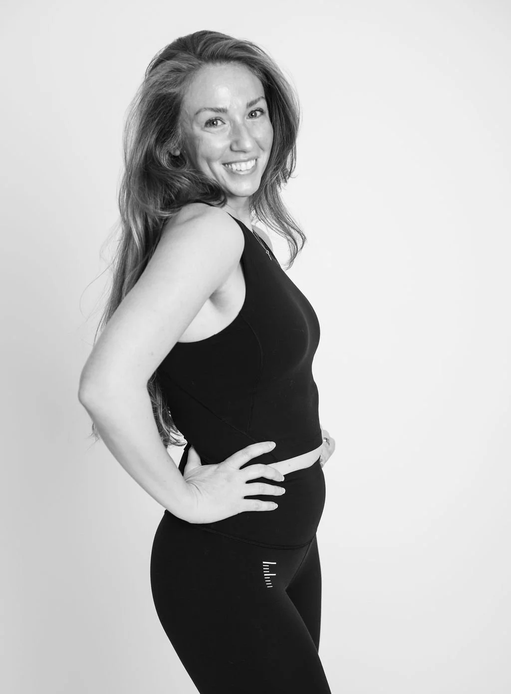 Black and white portrait of a smiling woman with long hair, wearing athletic clothing, standing with her hand on her hip against a plain background.