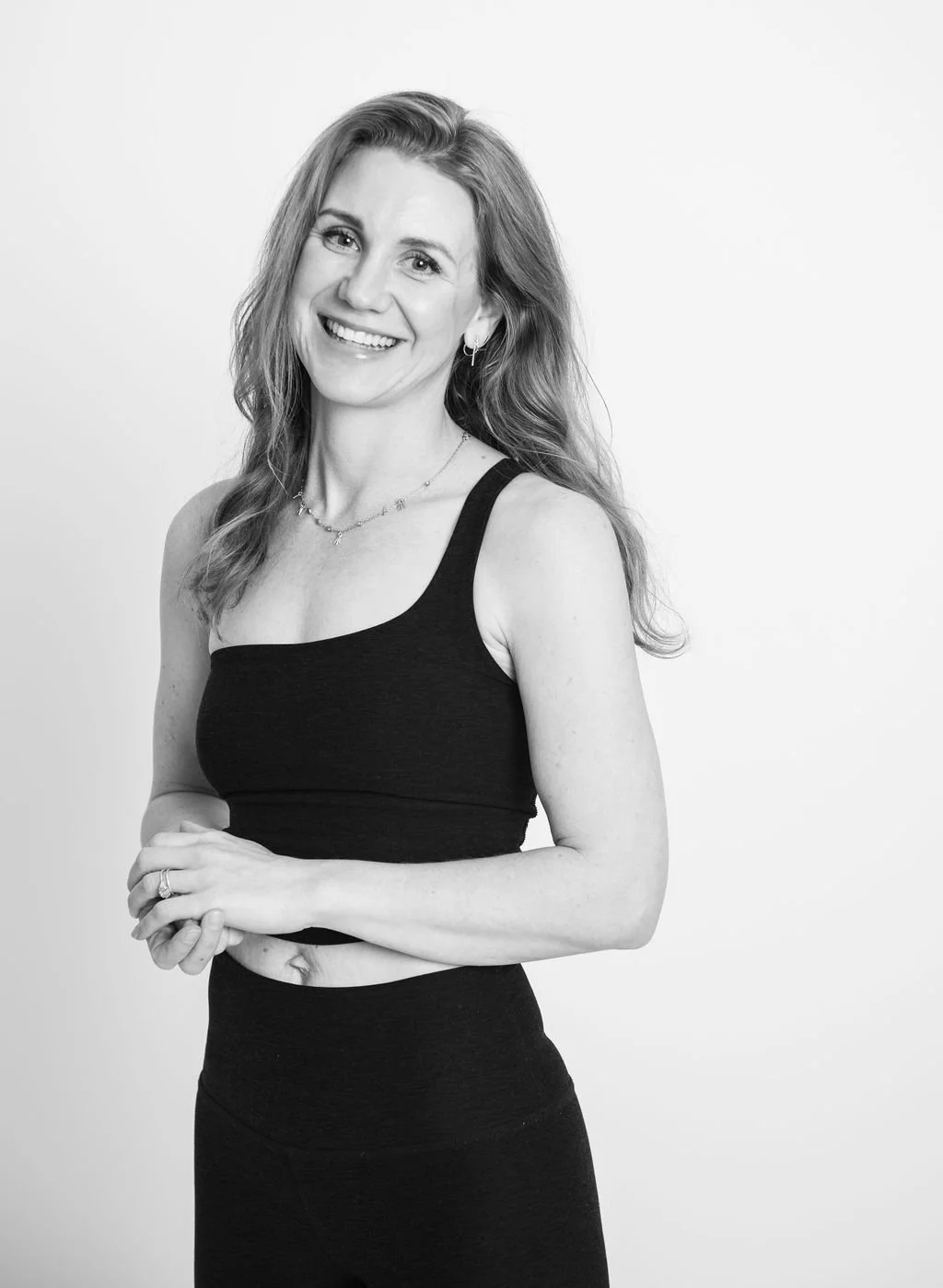 Black and white portrait of a smiling woman with long wavy hair wearing a sleeveless black top and jewelry, standing against a plain light background.