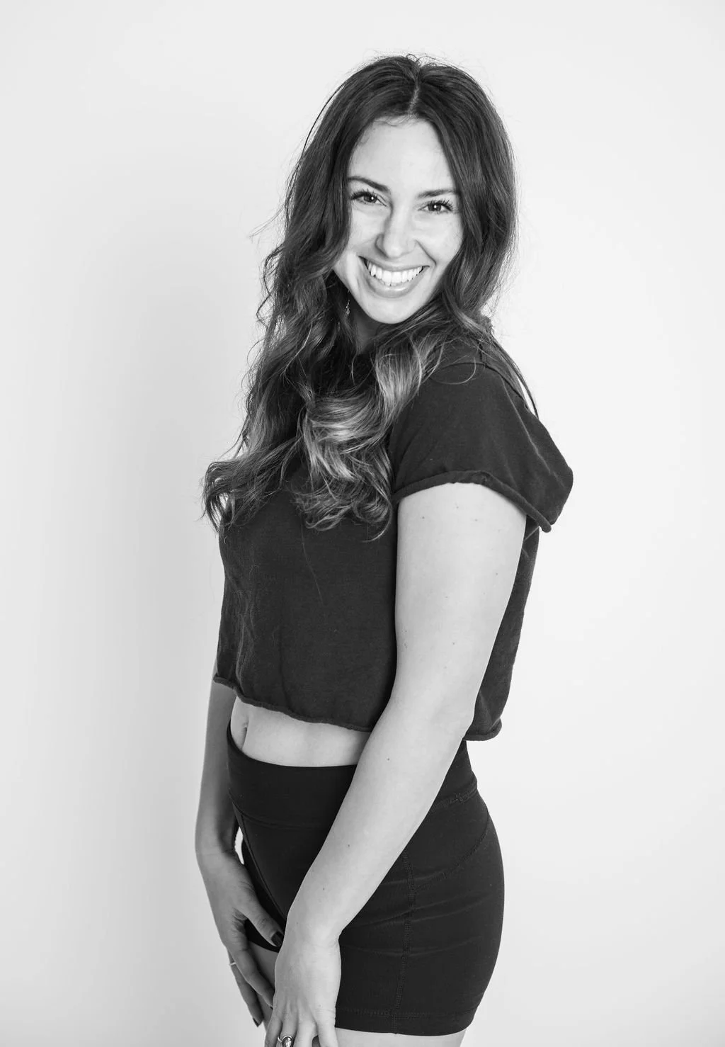 Black and white photo of a smiling young woman with long, wavy hair, wearing a cropped t-shirt and fitted shorts, posing against a plain background.