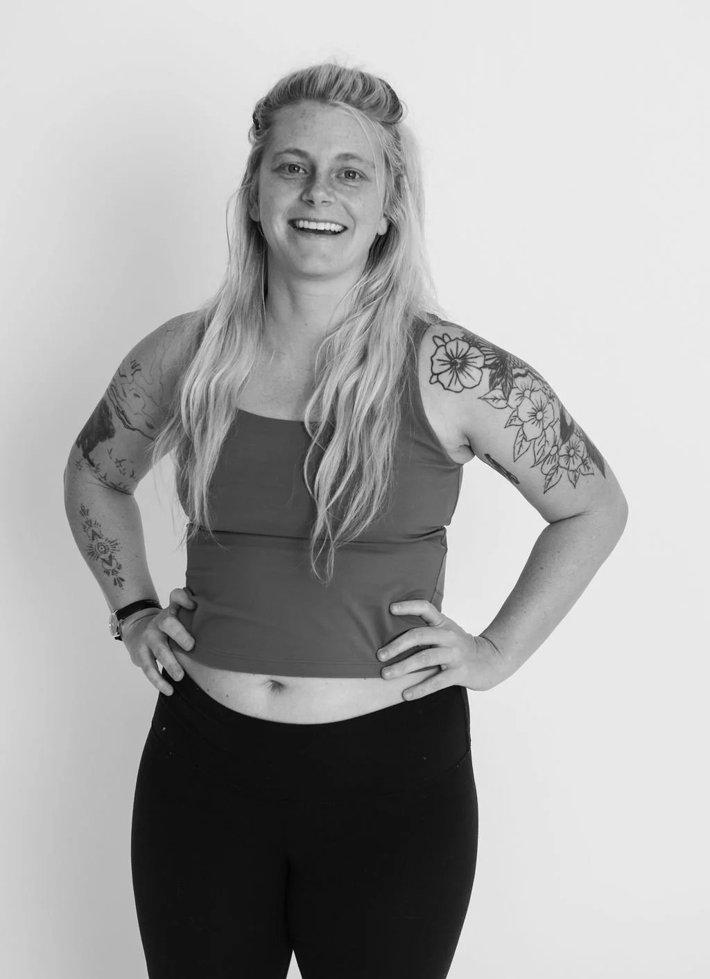 Smiling woman with long blonde hair, tattoos on her arms, wearing a sleeveless top and leggings, standing against a plain background.