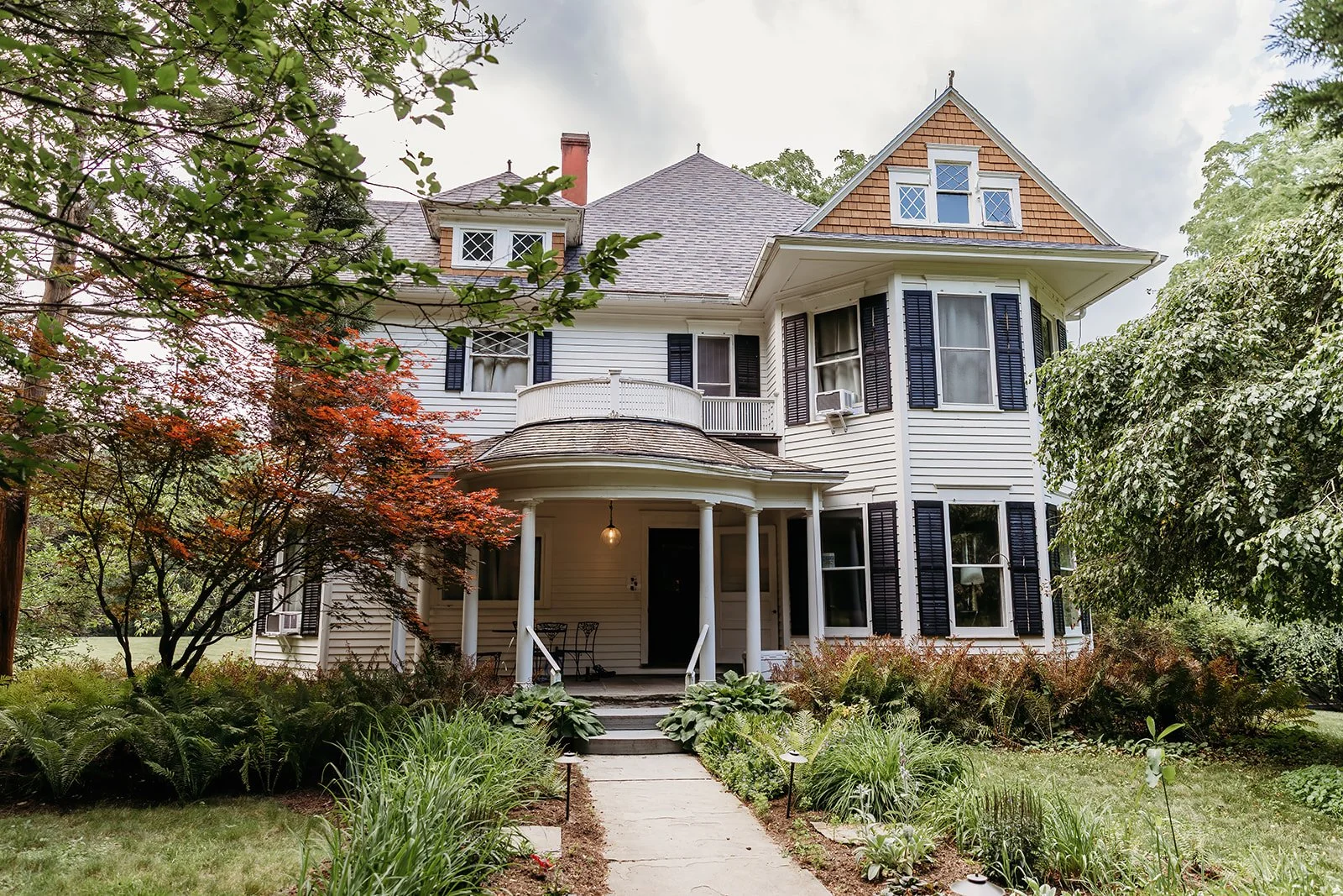 A large, multi-story Victorian style house with white wooden siding, black shutters, and a front porch with columns. The house has multiple windows, some with air conditioning units, and a distinctive gabled roof with shingles. Surrounding the house
