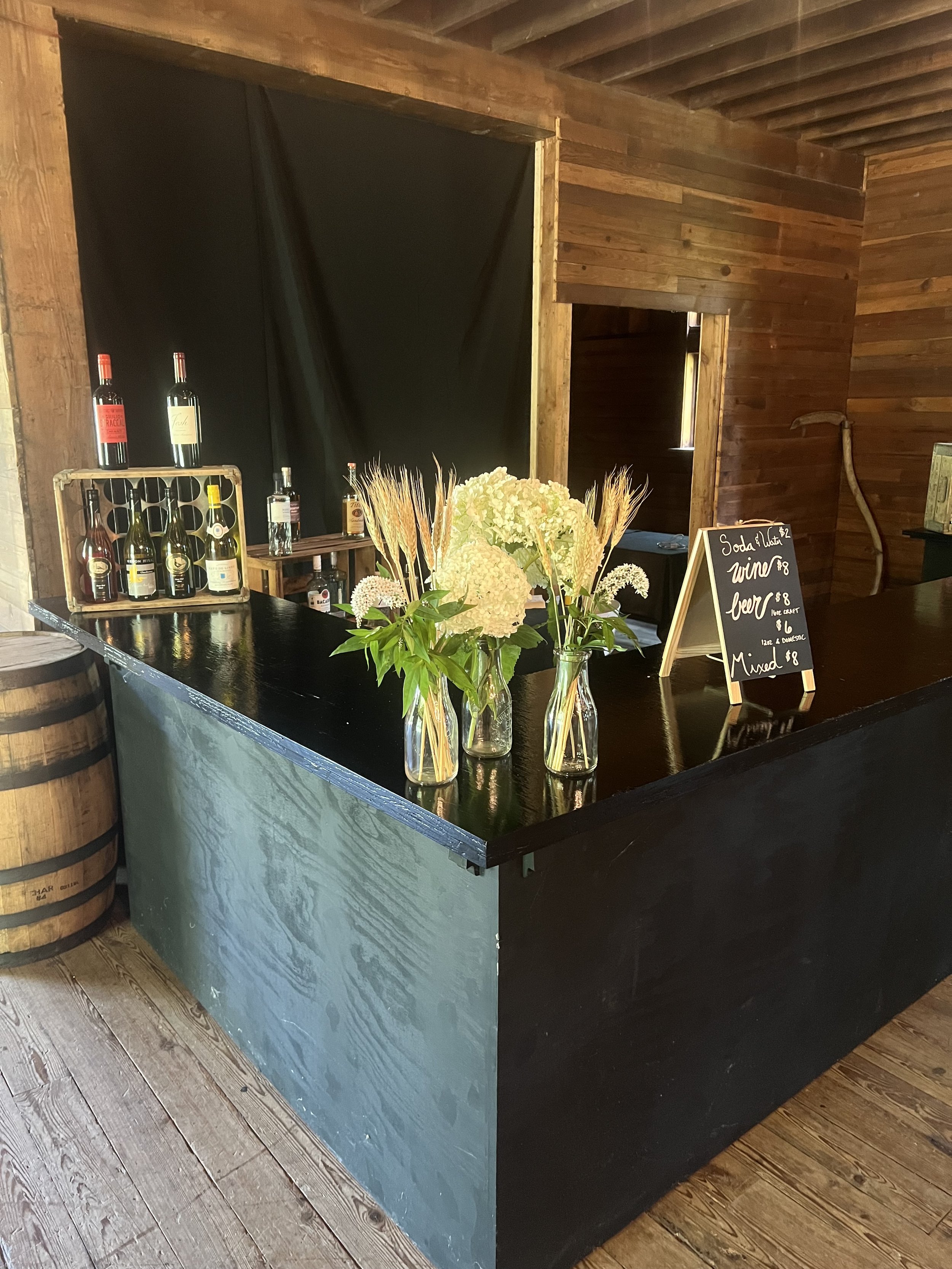 Rustic bar setup with a black counter, wooden wall backdrop, bottles of liquor on the left, floral arrangements in glass vases, and a chalkboard menu listing beverages like wine and beer. A barrel is partially visible next to the bar.