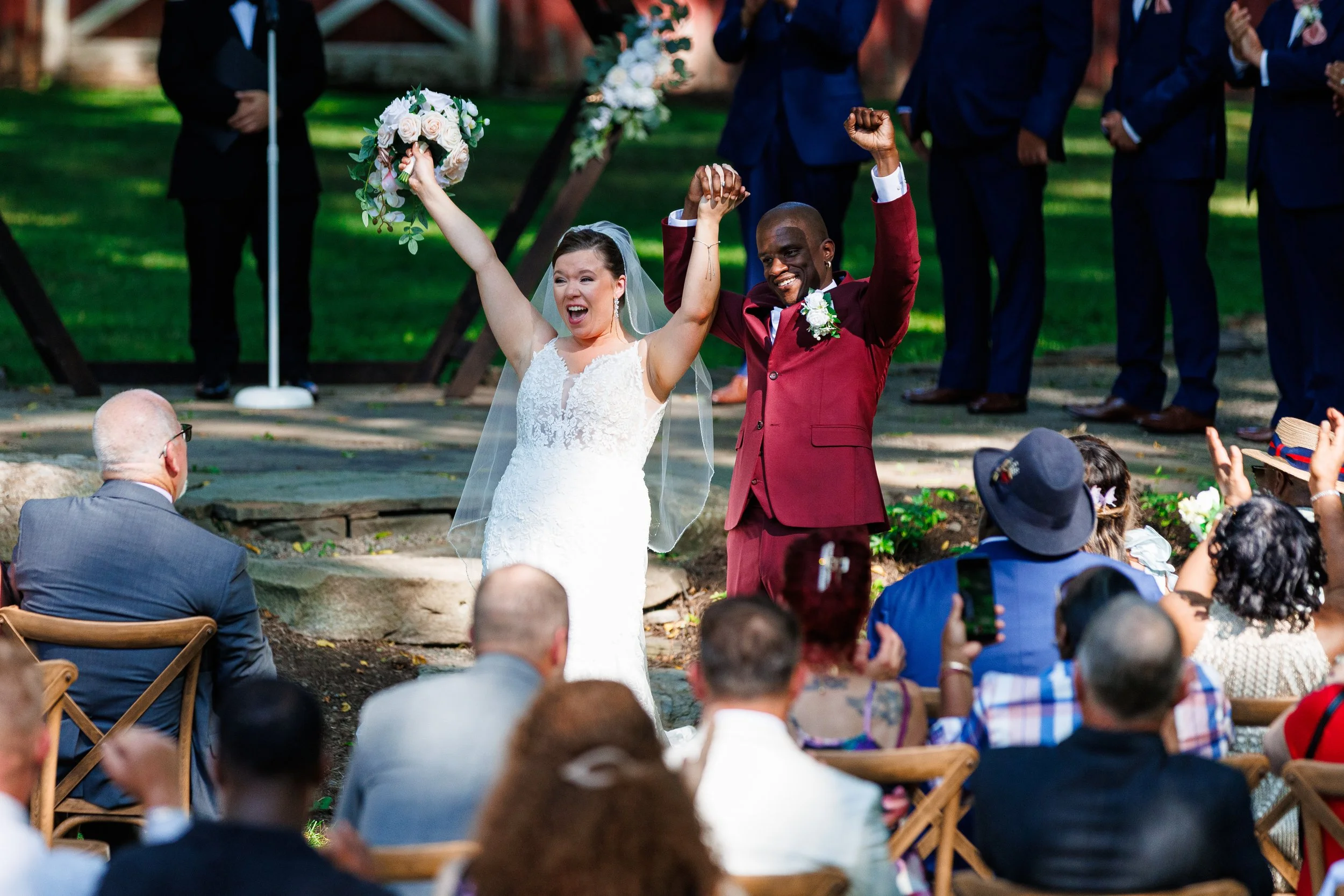 Bride and groom celebrating at their wedding ceremony outdoors, holding hands up in victory, with wedding guests applauding in the background.