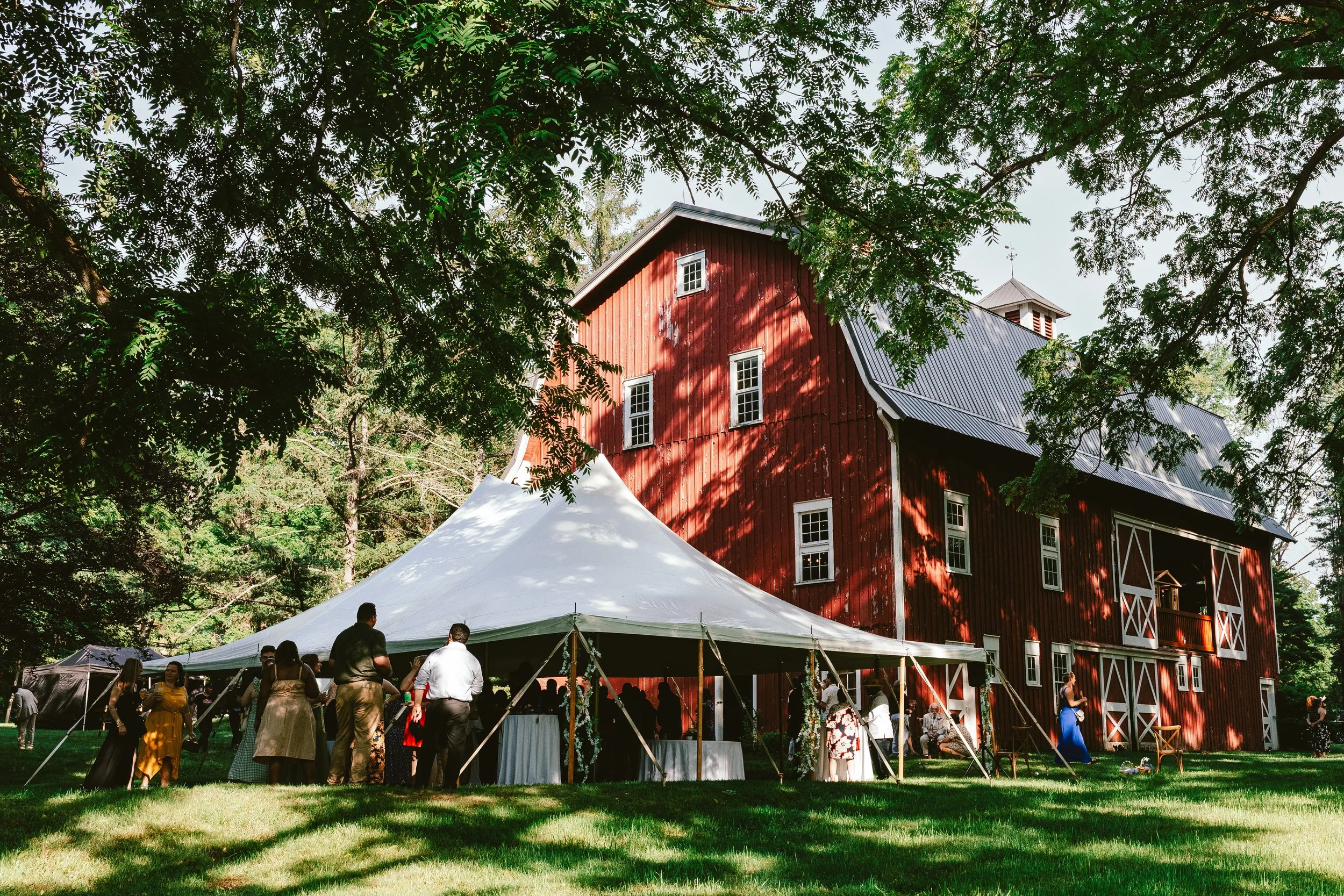 People gathering under a large white tent outside a red barn with white trim, surrounded by green trees.