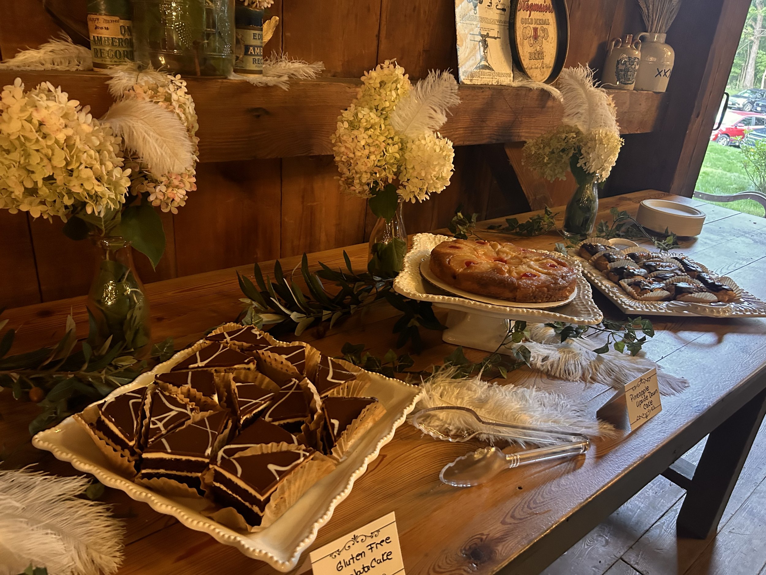 Dessert table with a variety of pastries including chocolate-layered bars, pineapple upside-down cake, and cookies, decorated with white hydrangeas and feathers.