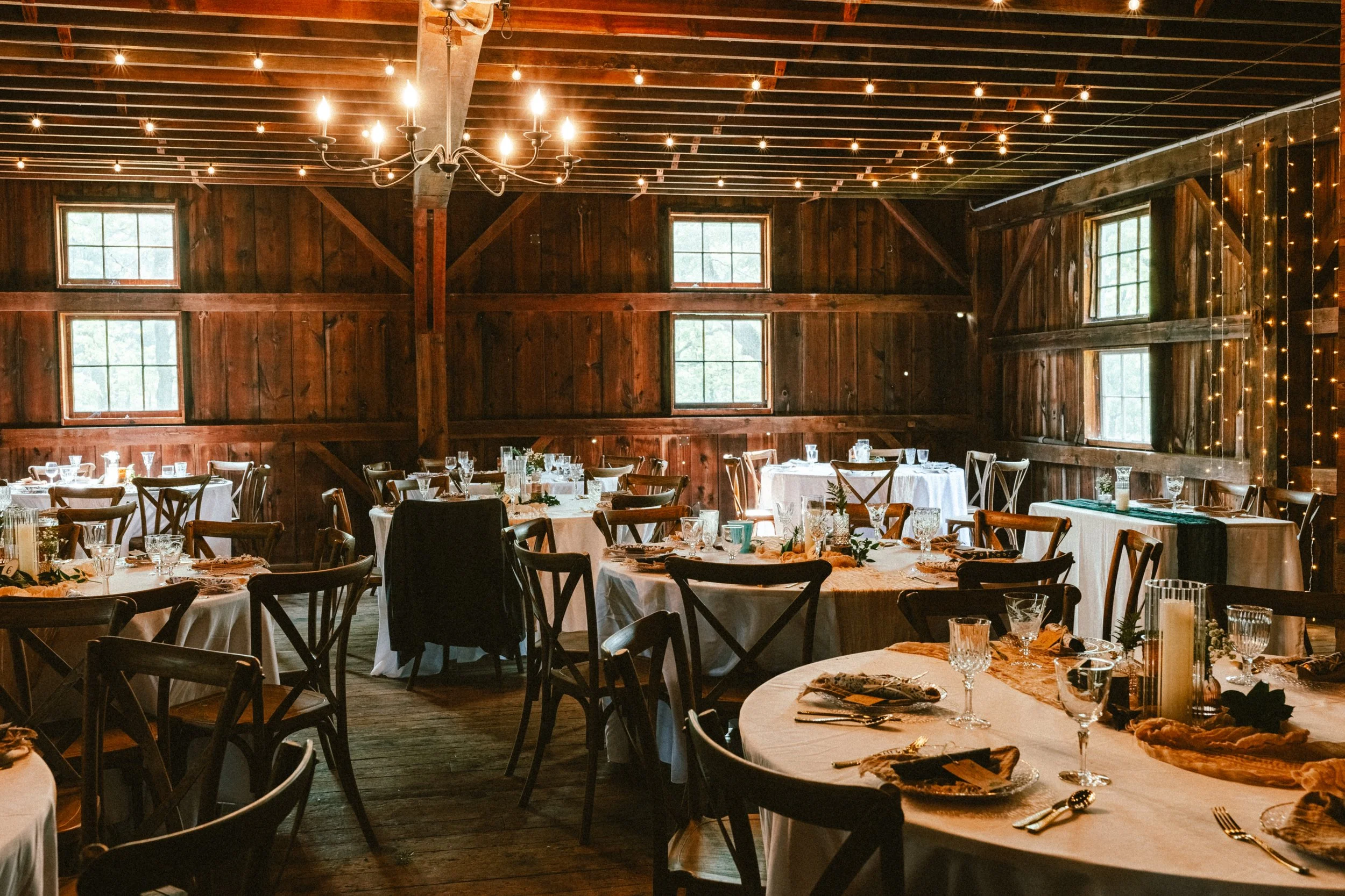 Rustic wooden banquet hall decorated for a celebration with round and rectangular tables set with plates, glasses, and napkins, illuminated by a chandelier and string lights, with windows letting in natural light.